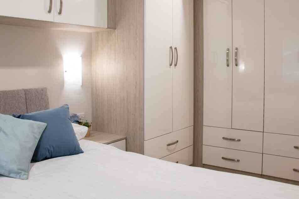 A cozy bedroom featuring a neatly made bed with light bedding and decorative pillows. There is a modern wardrobe with glossy white doors, a bedside table, and soft lighting illuminating the space, creating a welcoming atmosphere.