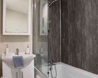 A modern bathroom featuring a bathtub with a glass shower enclosure. There is a sleek sink with a mirror above it, along with minimalist soap dispensers. The walls have a textured dark paneling, creating a contemporary look.