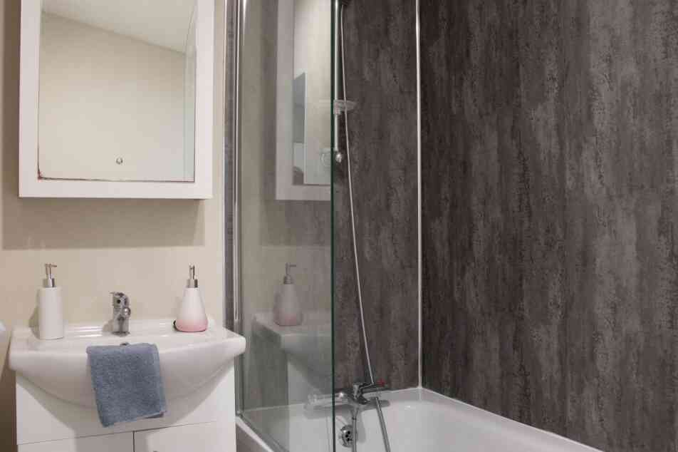 A modern bathroom featuring a bathtub with a glass shower enclosure. There is a sleek sink with a mirror above it, along with minimalist soap dispensers. The walls have a textured dark paneling, creating a contemporary look.