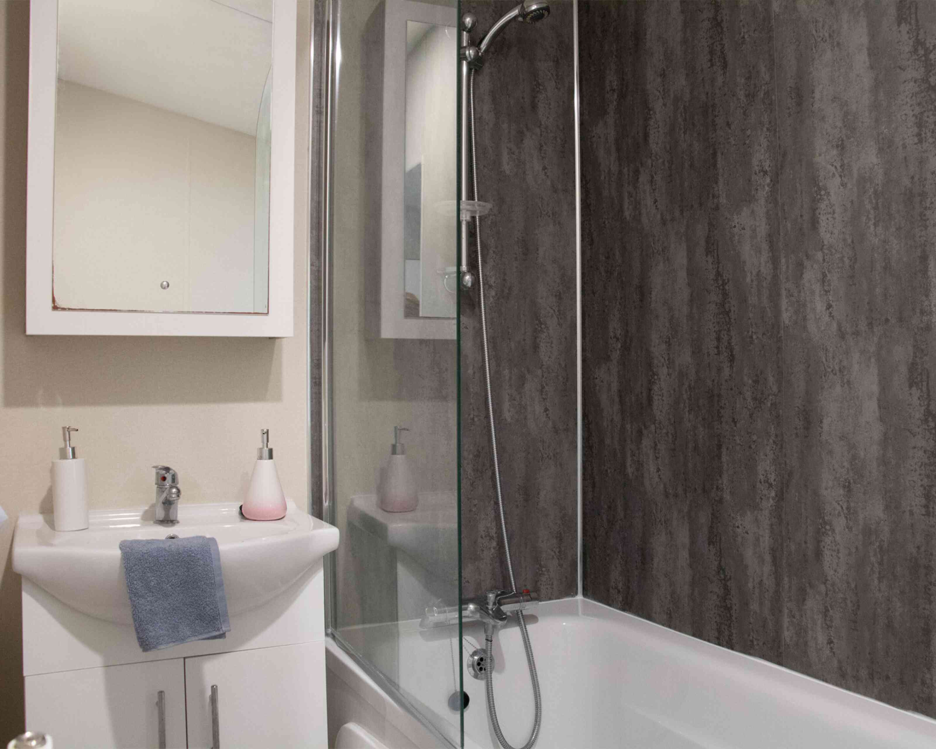 A modern bathroom featuring a bathtub with a glass shower enclosure. There is a sleek sink with a mirror above it, along with minimalist soap dispensers. The walls have a textured dark paneling, creating a contemporary look.