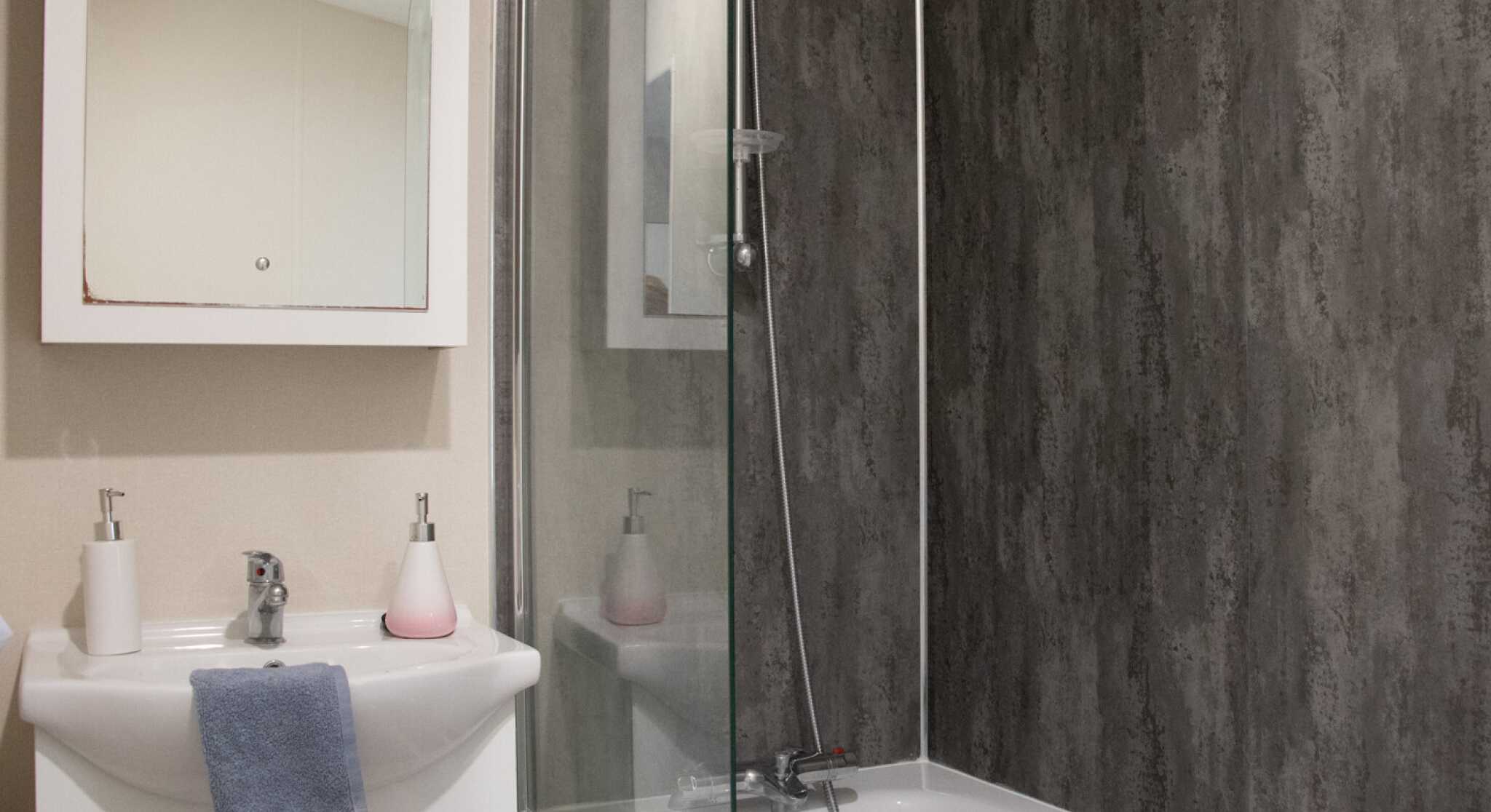A modern bathroom featuring a bathtub with a glass shower enclosure. There is a sleek sink with a mirror above it, along with minimalist soap dispensers. The walls have a textured dark paneling, creating a contemporary look.