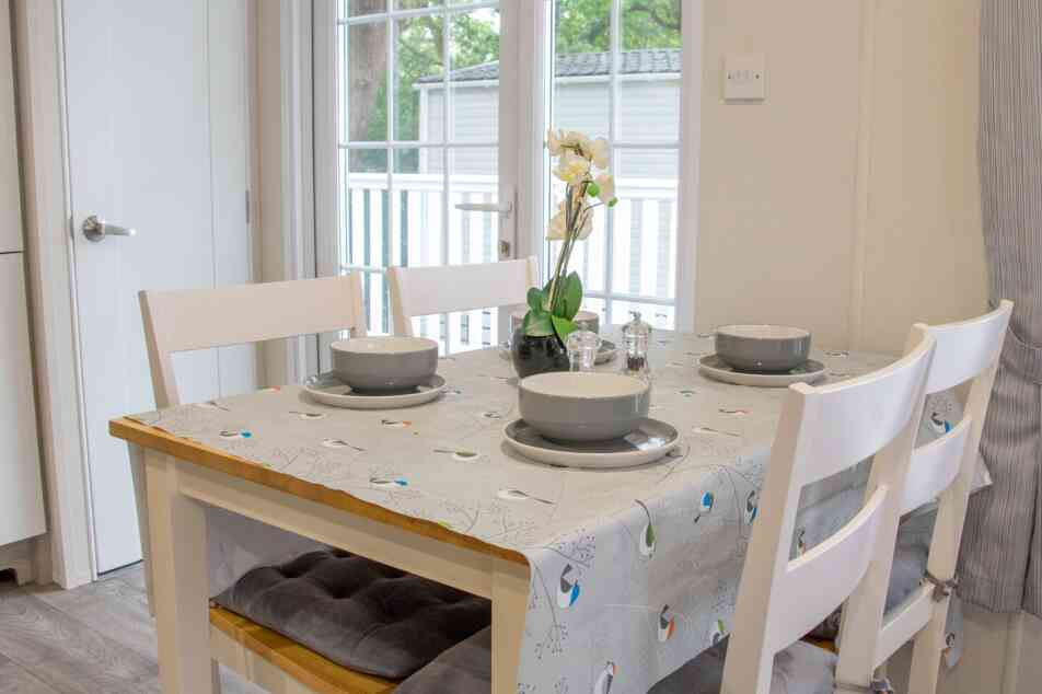 A wooden dining table with a gray tablecloth, set for four guests with plates and bowls. Chairs are arranged around the table. A small vase with an orchid and natural light comes in through a nearby window.