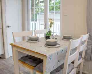 A wooden dining table with a gray tablecloth, set for four guests with plates and bowls. Chairs are arranged around the table. A small vase with an orchid and natural light comes in through a nearby window.