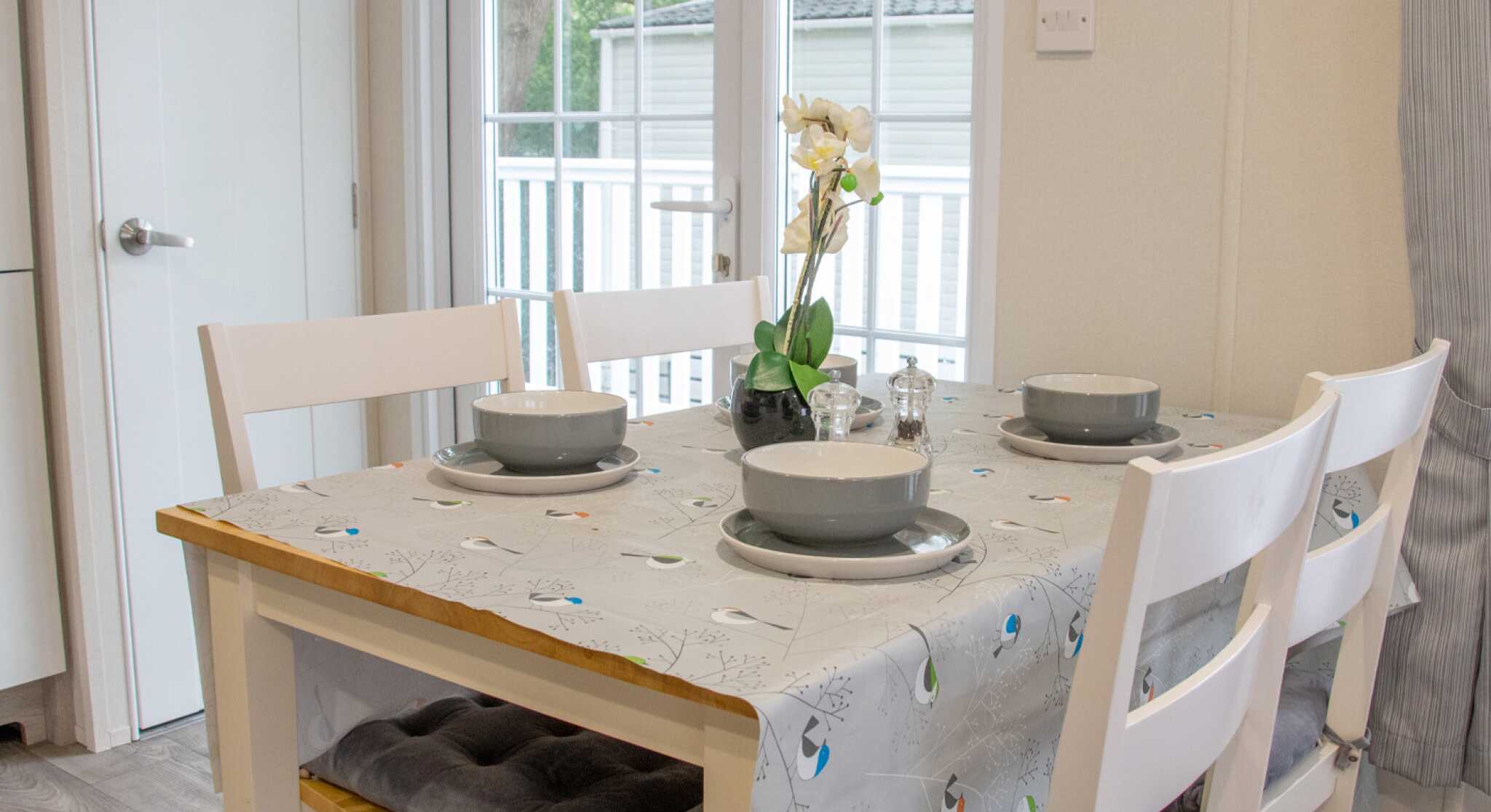 A wooden dining table with a gray tablecloth, set for four guests with plates and bowls. Chairs are arranged around the table. A small vase with an orchid and natural light comes in through a nearby window.