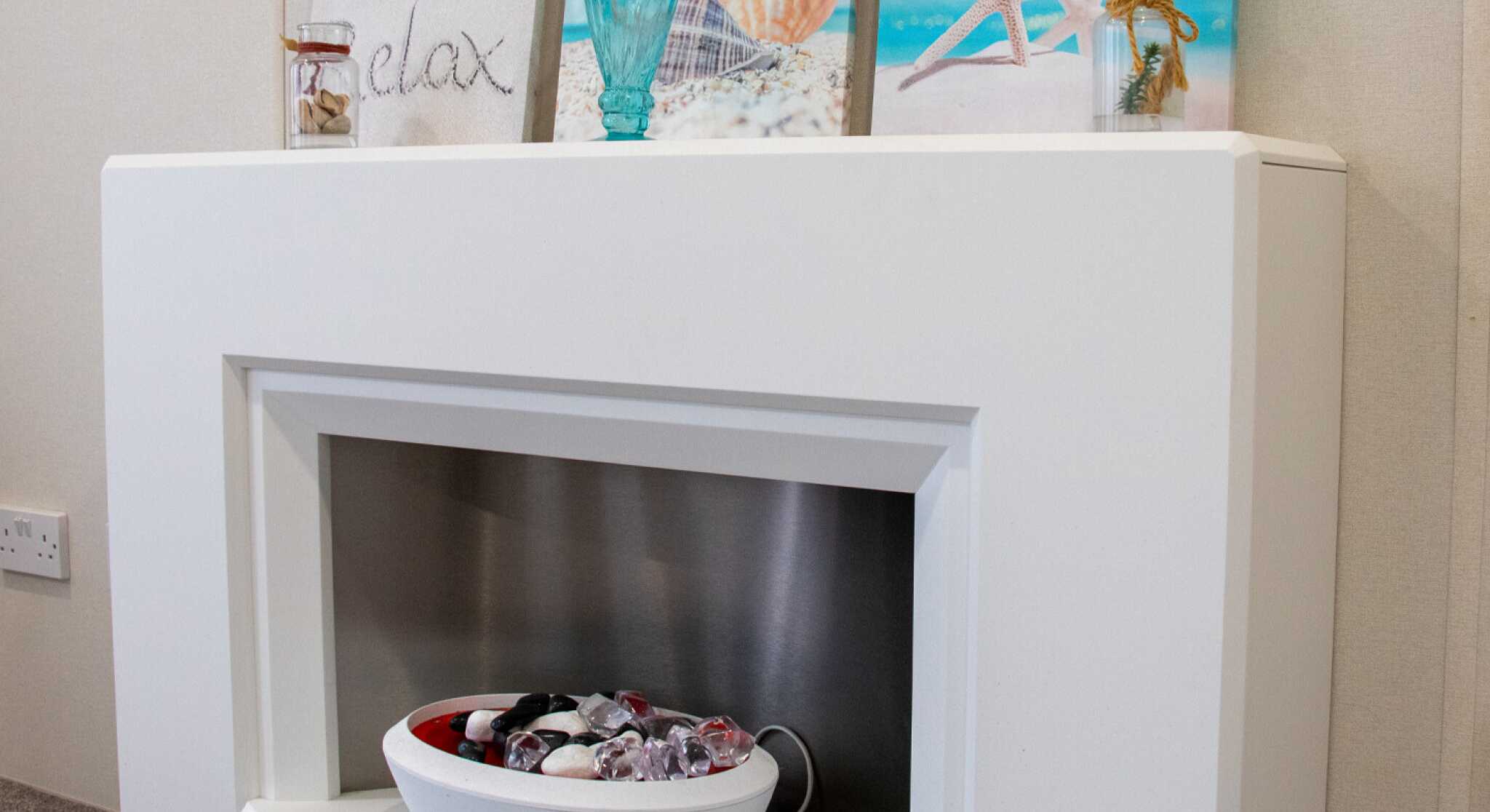 A modern white fireplace with a stainless steel insert, featuring a decorative bowl of colorful stones. Above the fireplace are three framed beach-themed prints, including images of a seashell, starfish, and a turquoise decorative vase. The overall decor is light and serene, evoking a coastal atmosphere.