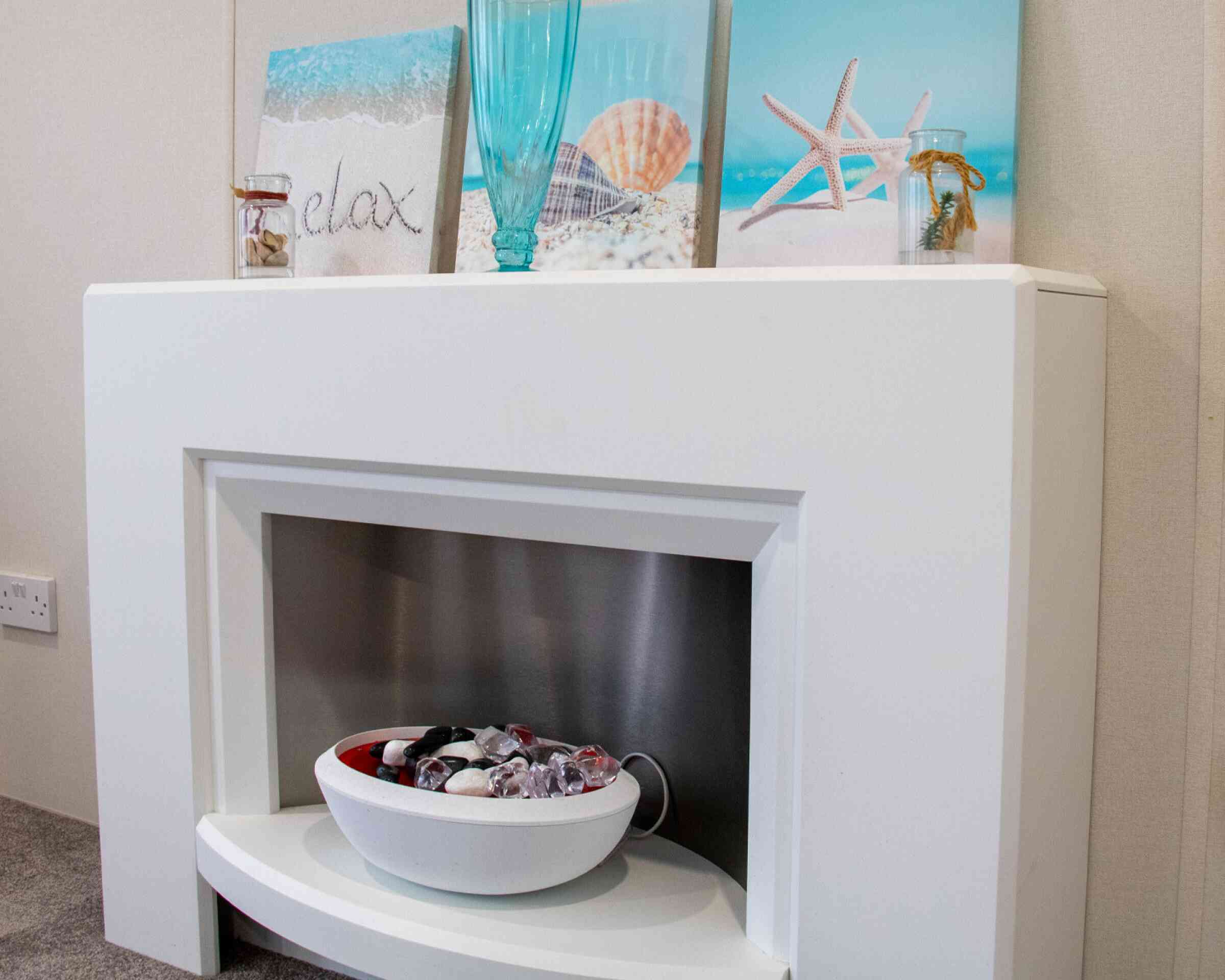 A modern white fireplace with a stainless steel insert, featuring a decorative bowl of colorful stones. Above the fireplace are three framed beach-themed prints, including images of a seashell, starfish, and a turquoise decorative vase. The overall decor is light and serene, evoking a coastal atmosphere.