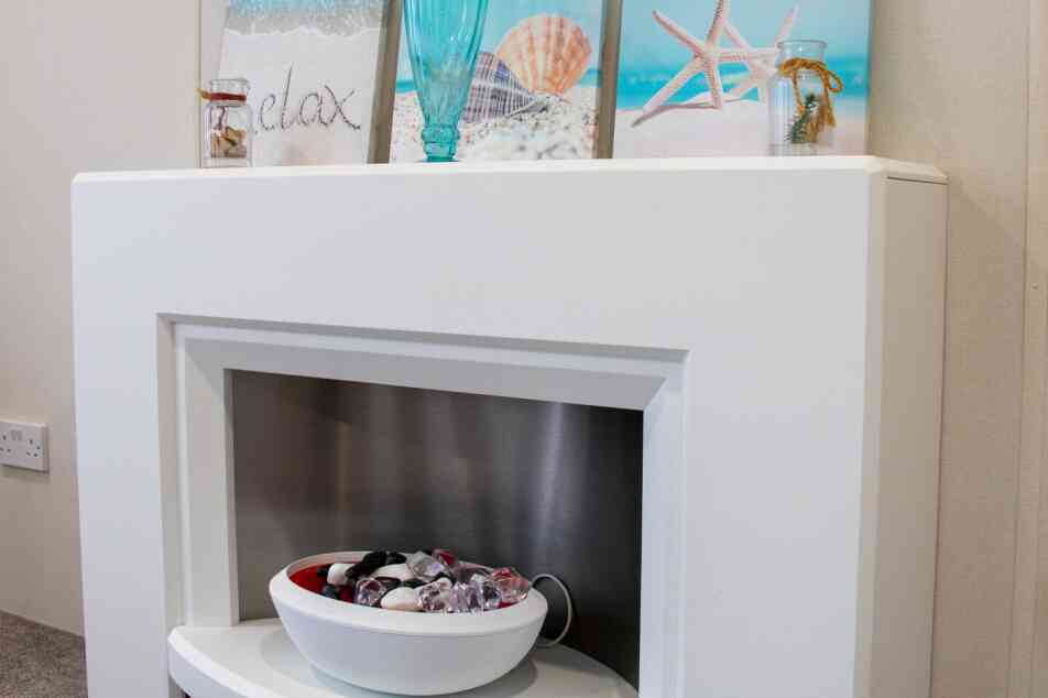 A modern white fireplace with a stainless steel insert, featuring a decorative bowl of colorful stones. Above the fireplace are three framed beach-themed prints, including images of a seashell, starfish, and a turquoise decorative vase. The overall decor is light and serene, evoking a coastal atmosphere.