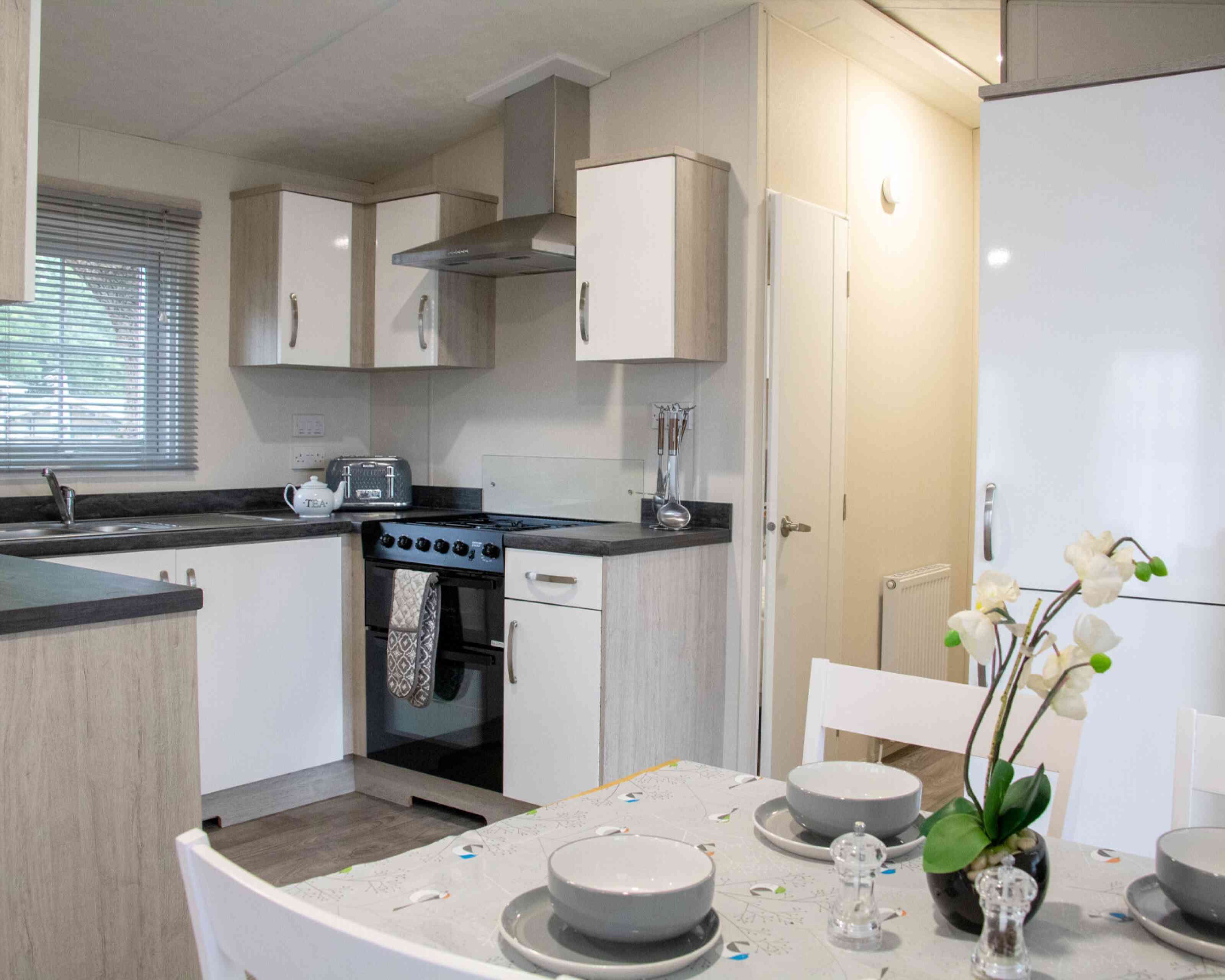 A modern kitchen featuring light wood cabinets, a gray countertop, and stainless steel appliances. The dining table is set with plates and glasses, and there is a vase with flowers on the table. Natural light streams through a window, illuminating the space.
