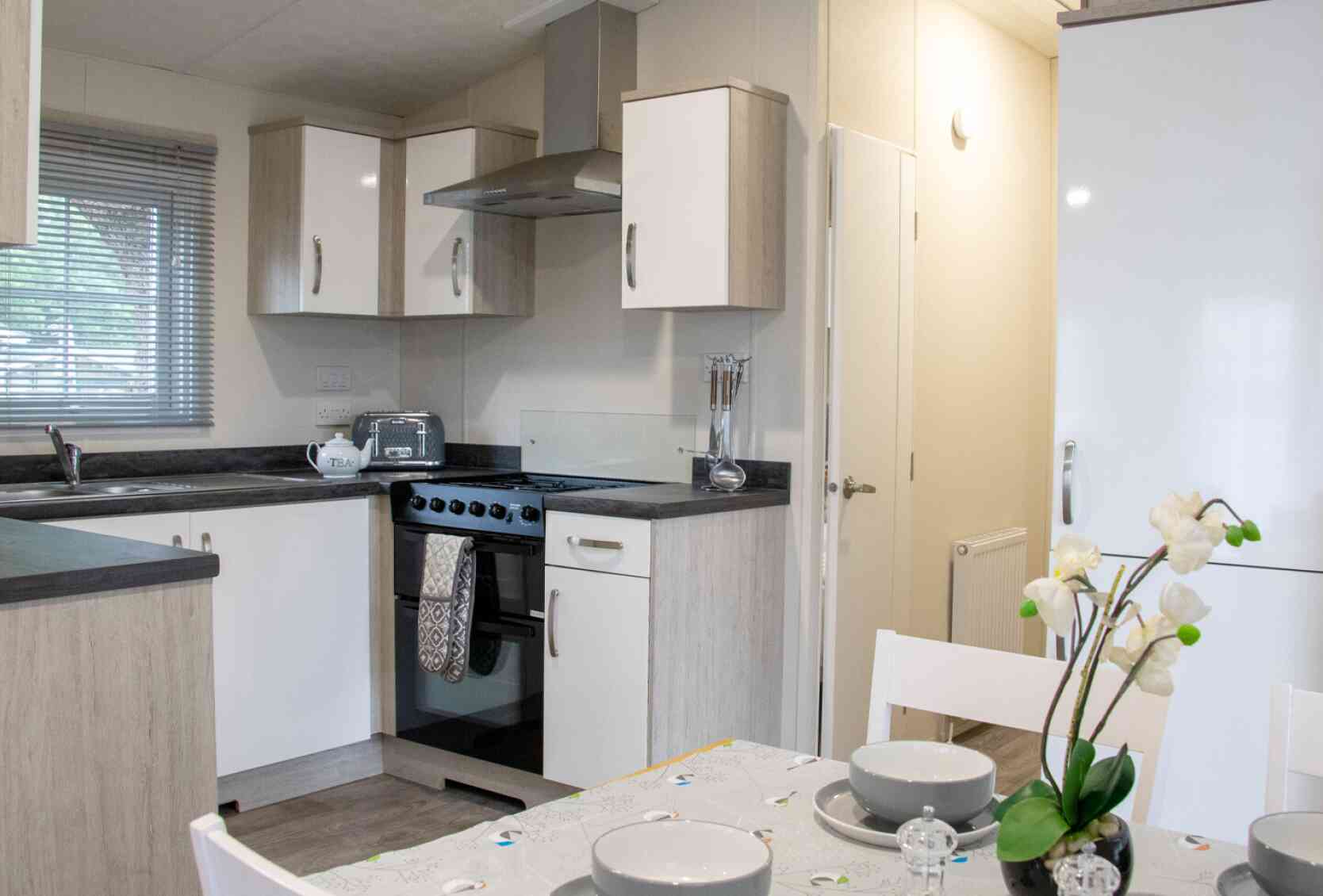A modern kitchen featuring light wood cabinets, a gray countertop, and stainless steel appliances. The dining table is set with plates and glasses, and there is a vase with flowers on the table. Natural light streams through a window, illuminating the space.