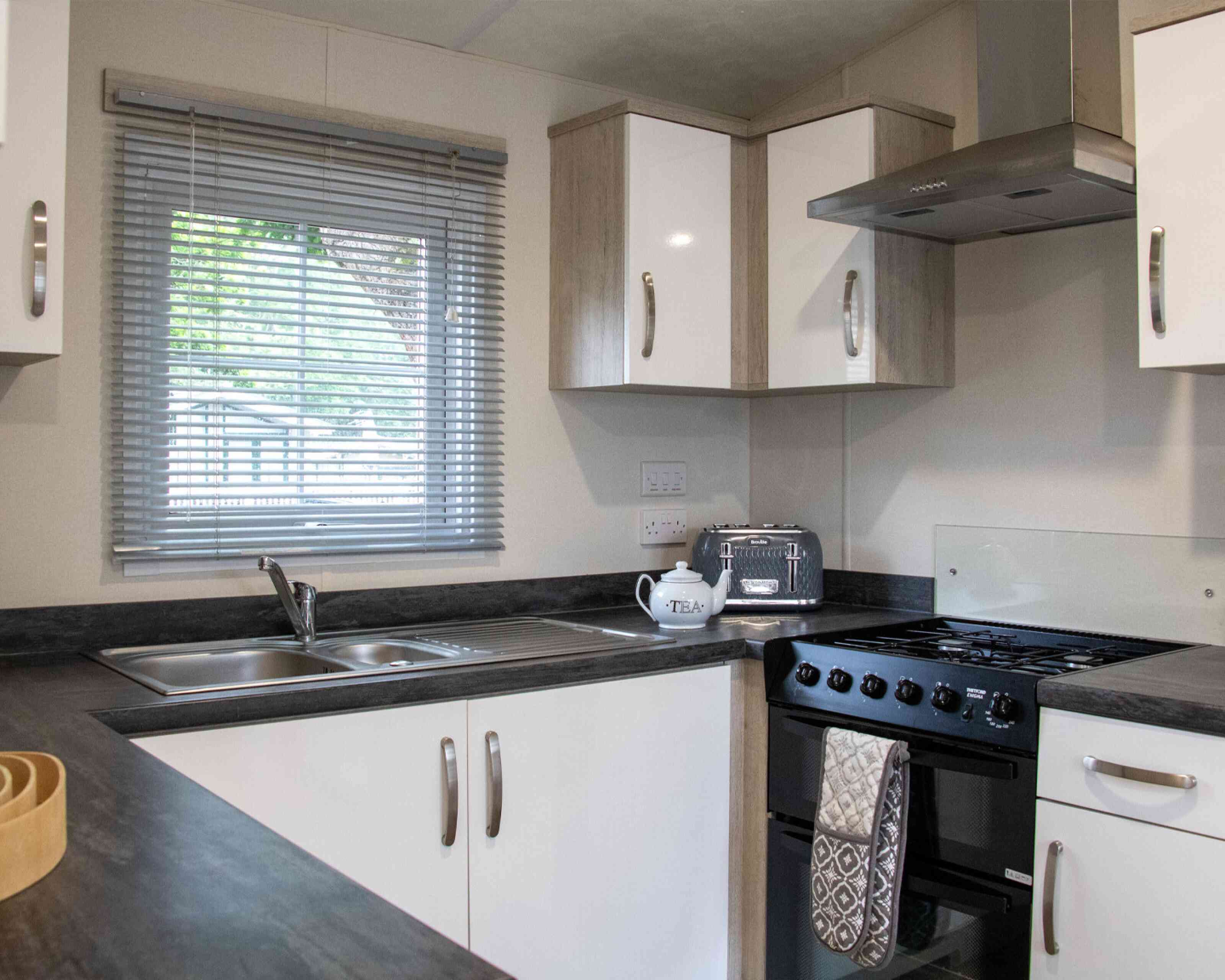 A modern kitchen featuring white cabinets, a black countertop, a stainless steel sink, and a gas stove. A window with blinds allows natural light to brighten the space. A decorative tea kettle is placed on the counter.