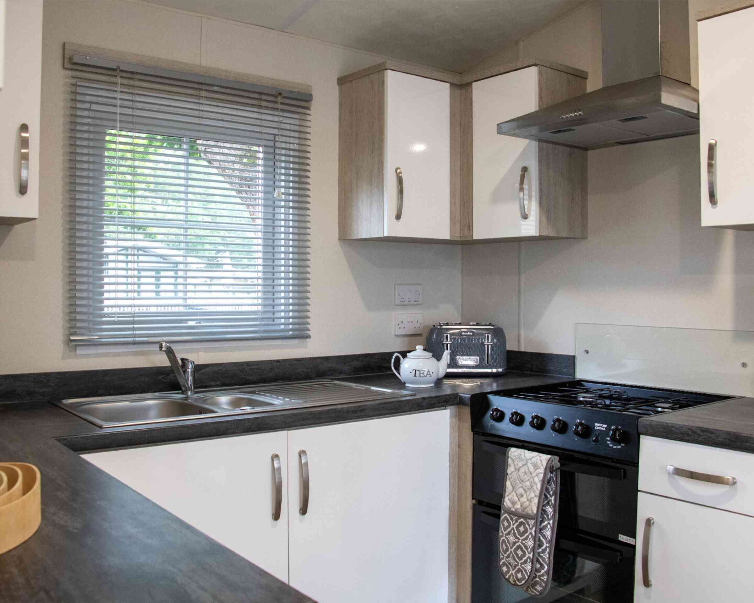 A modern kitchen featuring white cabinets, a black countertop, a stainless steel sink, and a gas stove. A window with blinds allows natural light to brighten the space. A decorative tea kettle is placed on the counter.