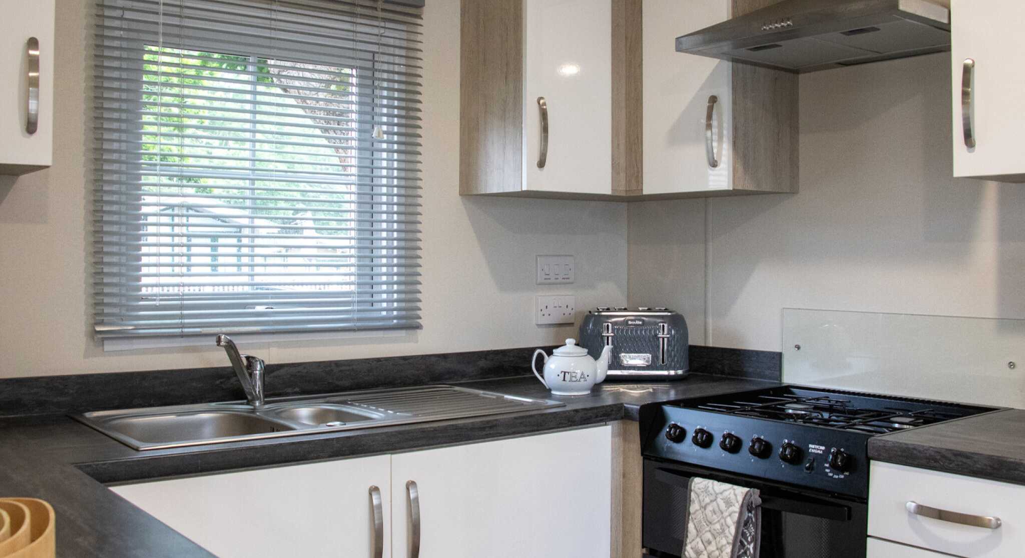 A modern kitchen featuring white cabinets, a black countertop, a stainless steel sink, and a gas stove. A window with blinds allows natural light to brighten the space. A decorative tea kettle is placed on the counter.