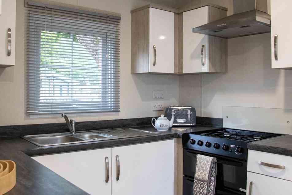 A modern kitchen featuring white cabinets, a black countertop, a stainless steel sink, and a gas stove. A window with blinds allows natural light to brighten the space. A decorative tea kettle is placed on the counter.