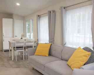 A cozy interior of a light-filled living space featuring a gray sofa with yellow cushions, a dining table with chairs, and large windows dressed with sheer curtains. A white cupboard is visible in the background.
