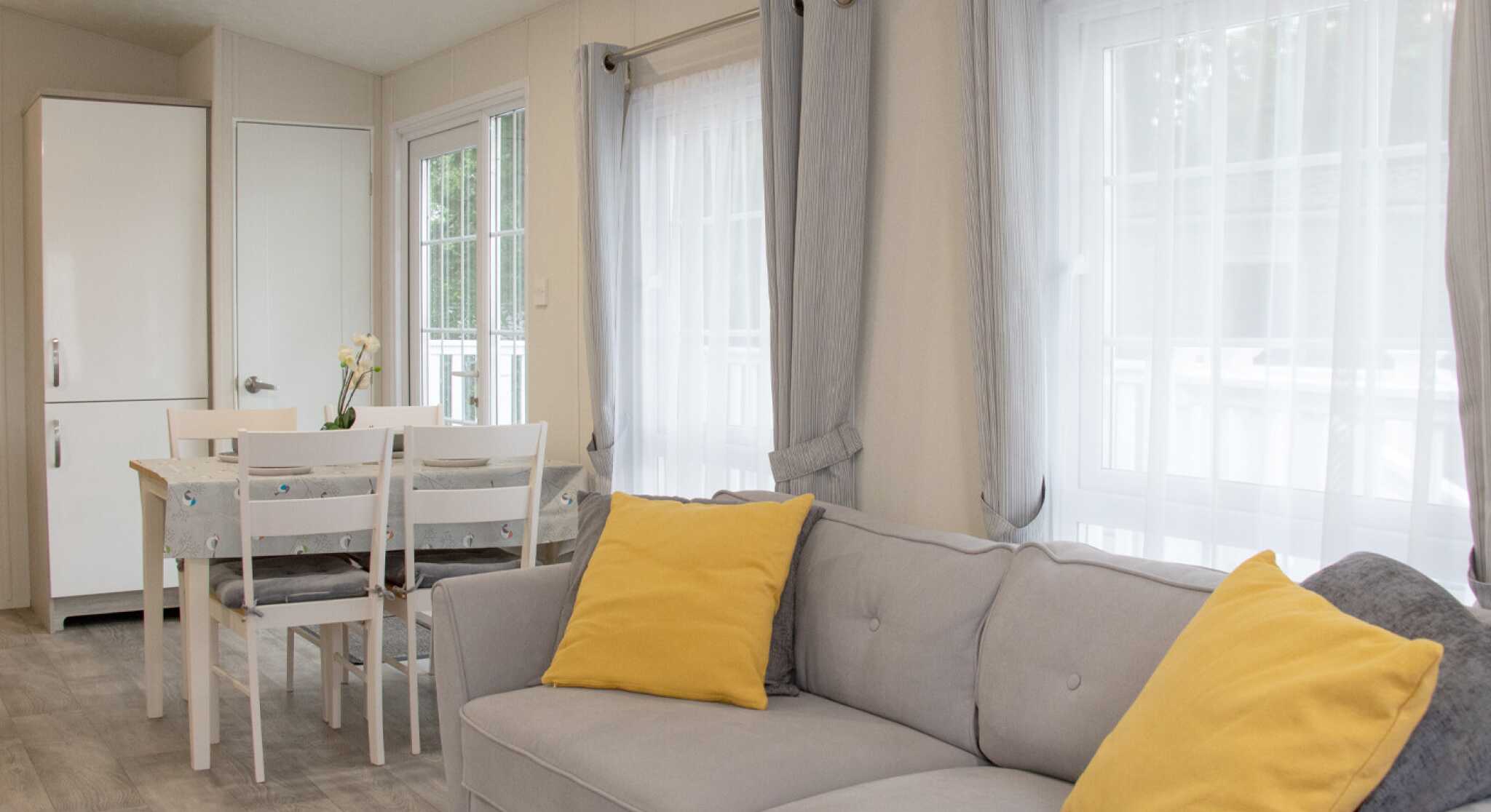 A cozy interior of a light-filled living space featuring a gray sofa with yellow cushions, a dining table with chairs, and large windows dressed with sheer curtains. A white cupboard is visible in the background.