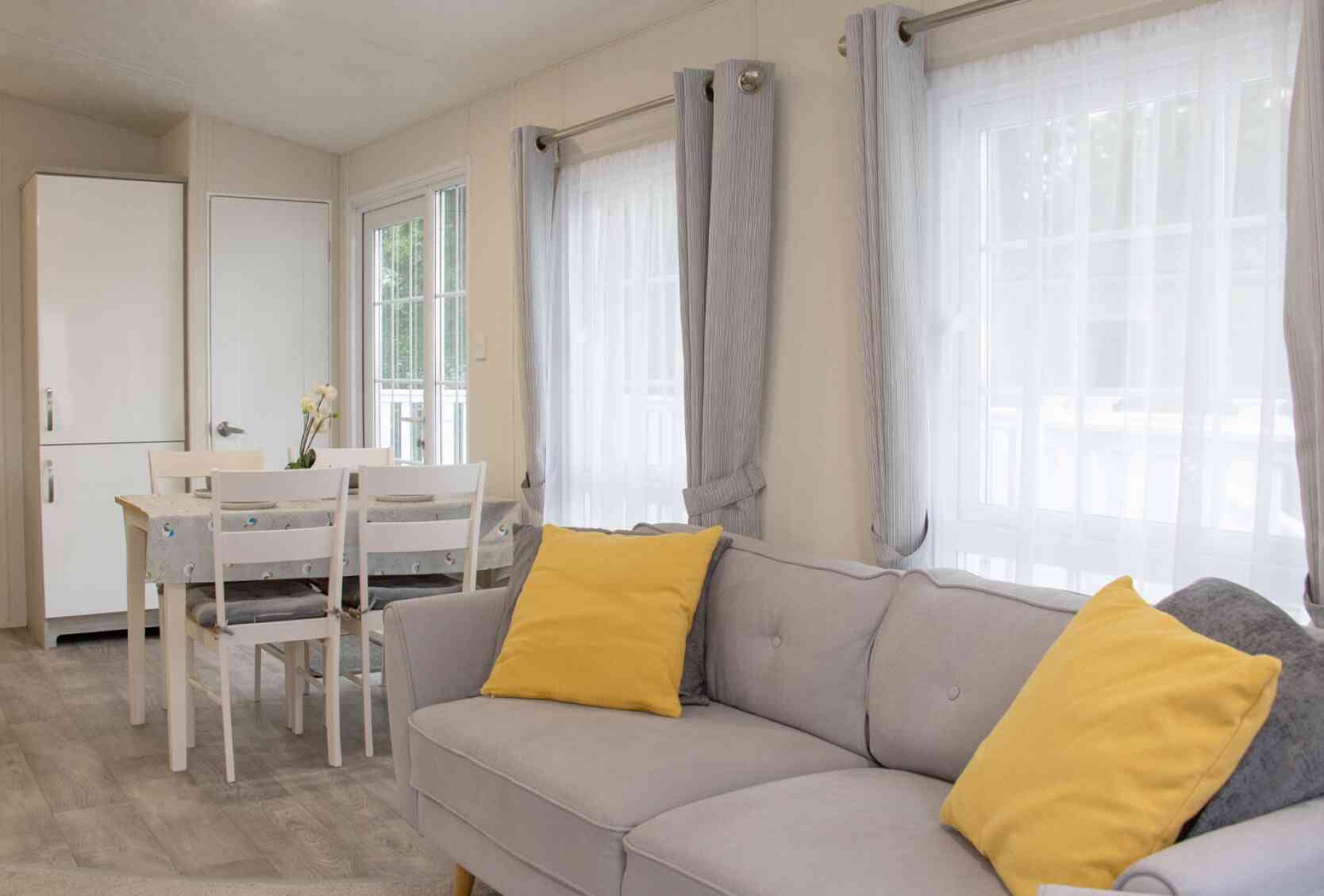 A cozy interior of a light-filled living space featuring a gray sofa with yellow cushions, a dining table with chairs, and large windows dressed with sheer curtains. A white cupboard is visible in the background.