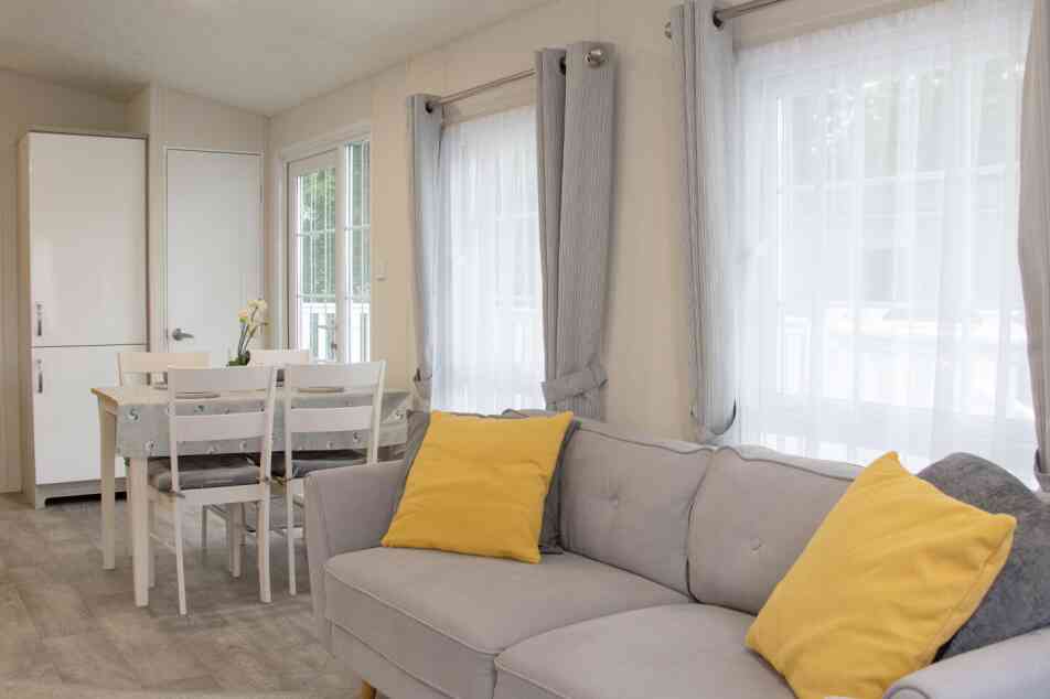 A cozy interior of a light-filled living space featuring a gray sofa with yellow cushions, a dining table with chairs, and large windows dressed with sheer curtains. A white cupboard is visible in the background.