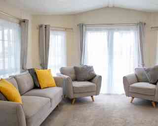 A cozy living area featuring light grey sofas with yellow and grey cushions. Large windows draped with sheer white curtains let in natural light, illuminating the carpeted floor.