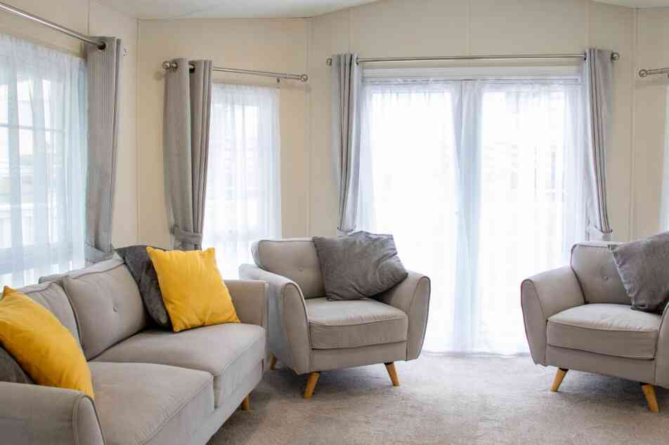 A cozy living area featuring light grey sofas with yellow and grey cushions. Large windows draped with sheer white curtains let in natural light, illuminating the carpeted floor.