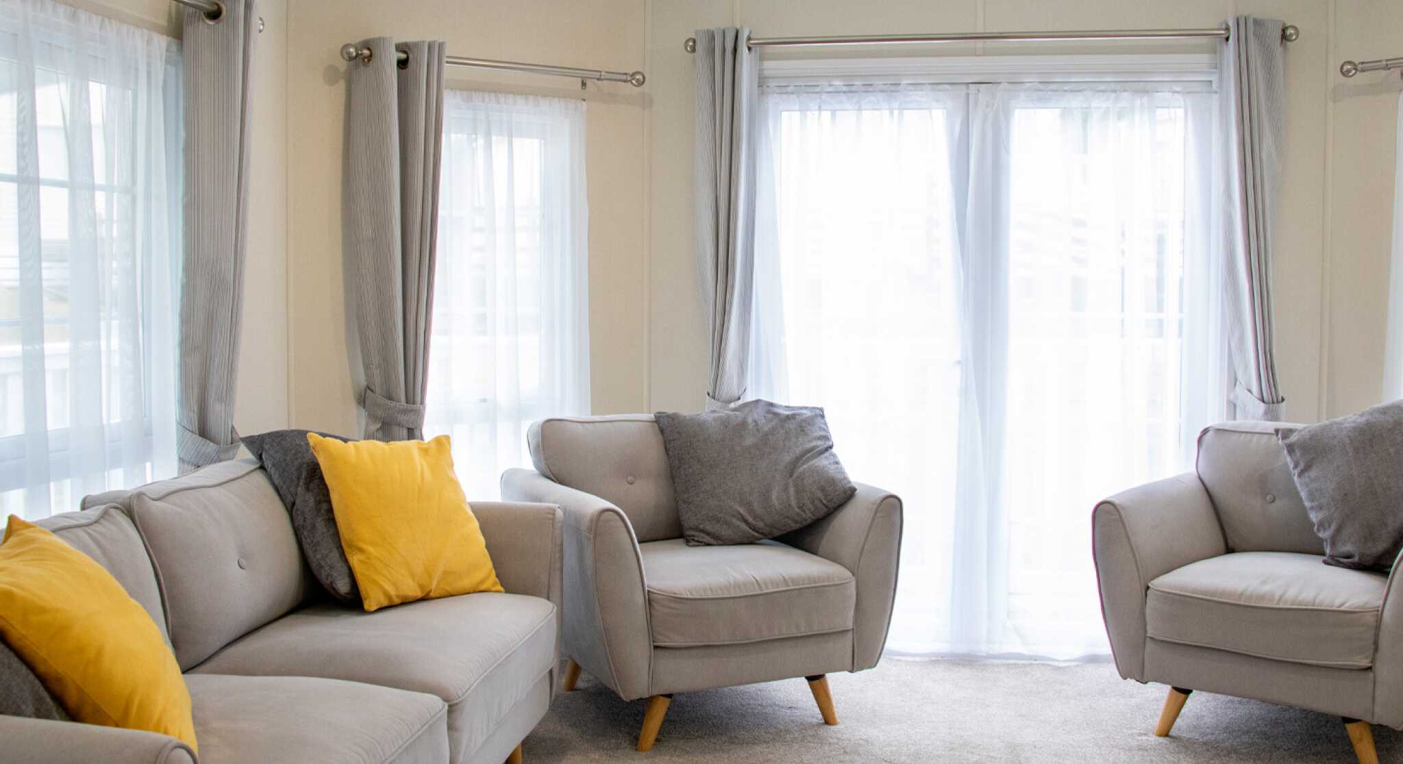 A cozy living area featuring light grey sofas with yellow and grey cushions. Large windows draped with sheer white curtains let in natural light, illuminating the carpeted floor.