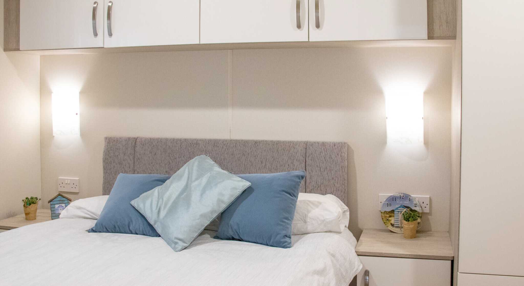 A cozy bedroom featuring a light gray upholstered headboard with blue decorative pillows. A white duvet covers the bed, and there are small potted plants on the nightstands. Wall-mounted lamps provide soft lighting above the bed, accompanied by fitted cabinets above.