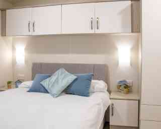 A cozy bedroom featuring a light gray upholstered headboard with blue decorative pillows. A white duvet covers the bed, and there are small potted plants on the nightstands. Wall-mounted lamps provide soft lighting above the bed, accompanied by fitted cabinets above.
