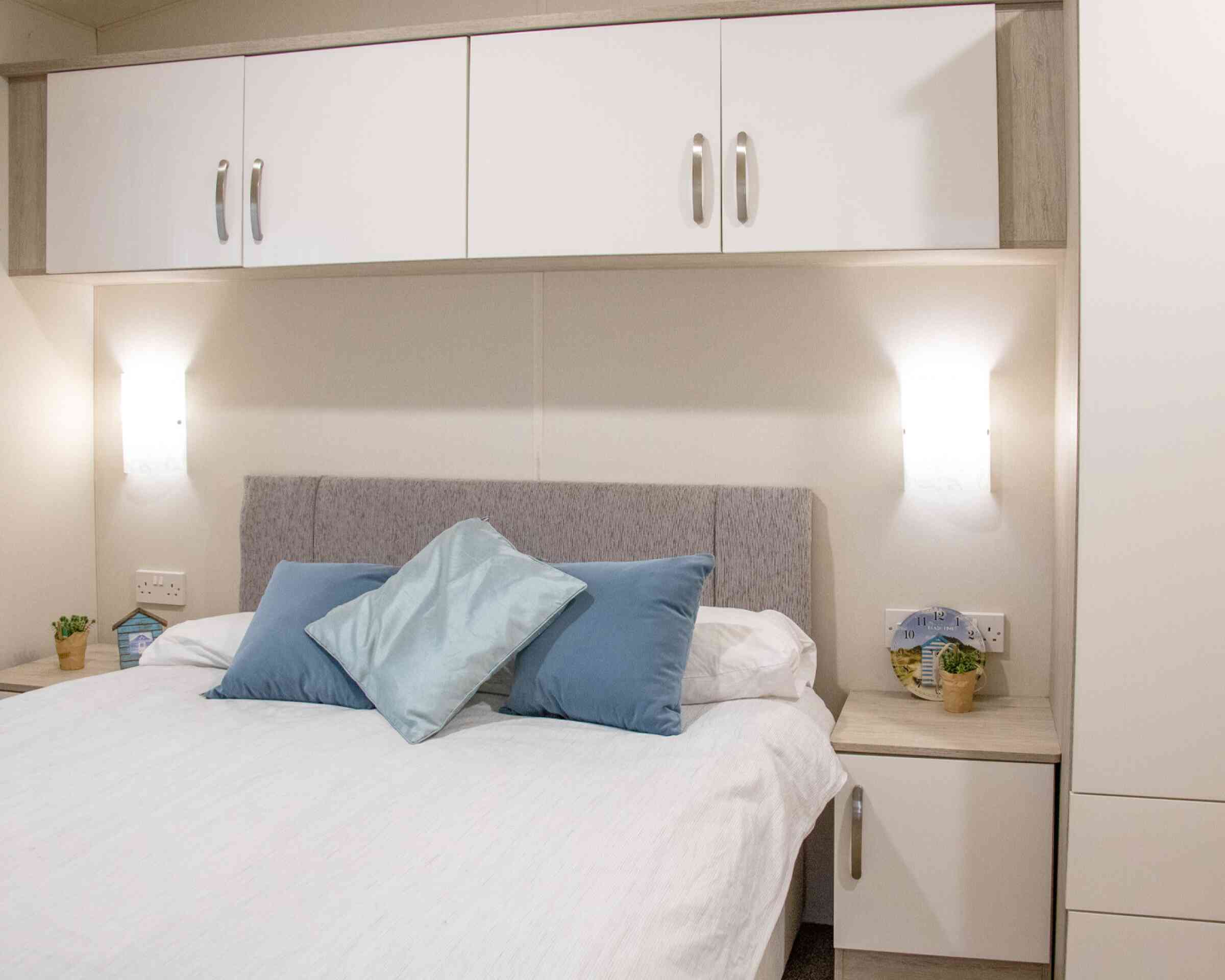 A cozy bedroom featuring a light gray upholstered headboard with blue decorative pillows. A white duvet covers the bed, and there are small potted plants on the nightstands. Wall-mounted lamps provide soft lighting above the bed, accompanied by fitted cabinets above.