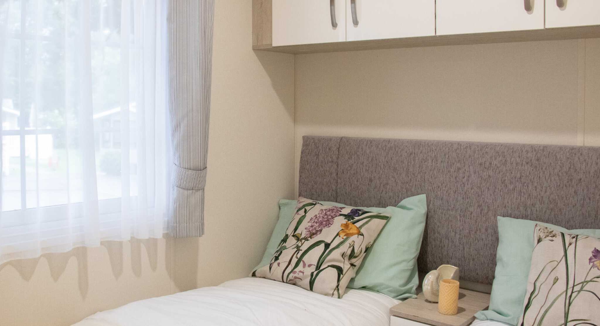 A cozy bedroom featuring two twin beds with light blue and floral-patterned pillows. There is a neutral-colored headboard and small bedside tables with decorative items. Natural light filters through a sheer window curtain.