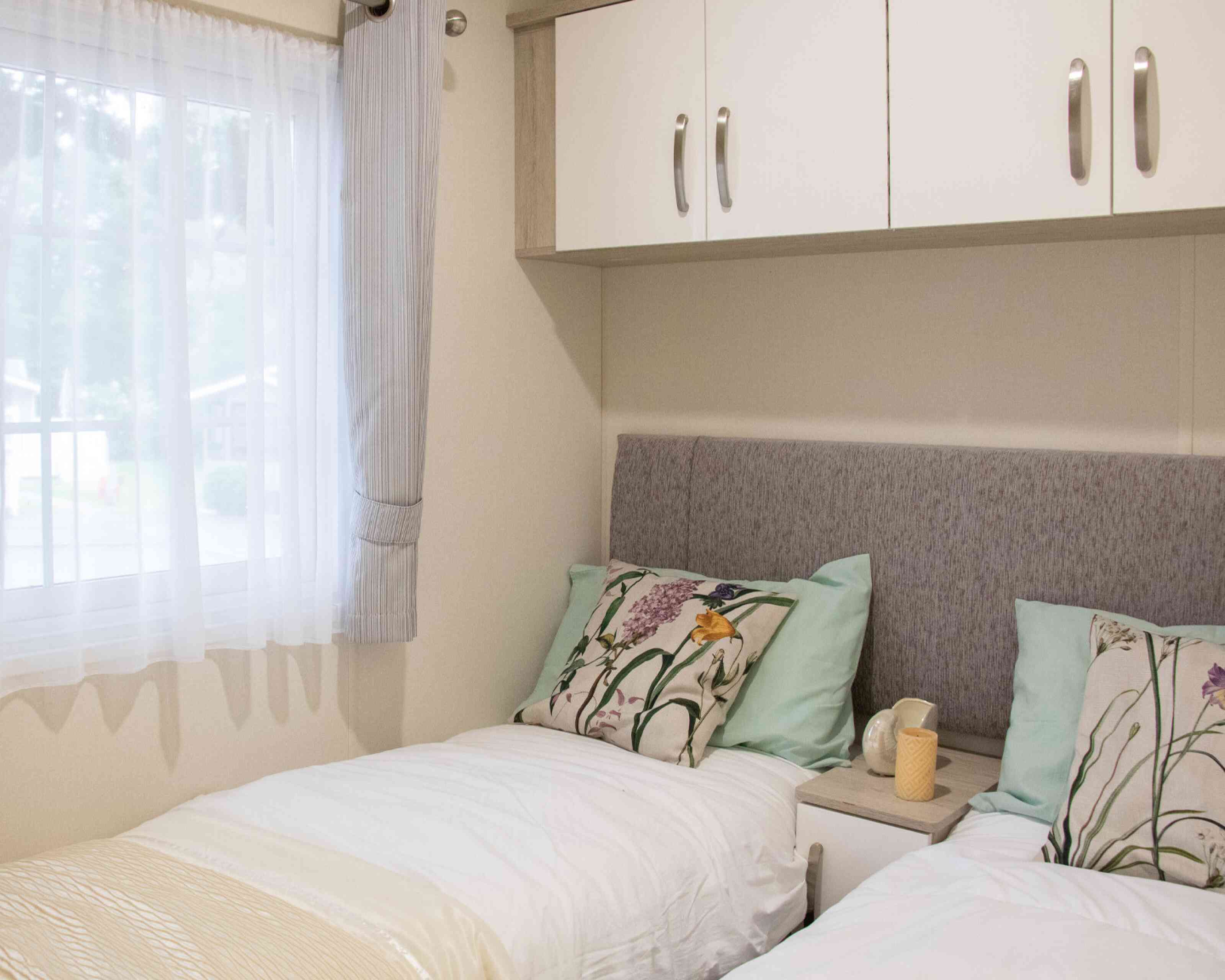 A cozy bedroom featuring two twin beds with light blue and floral-patterned pillows. There is a neutral-colored headboard and small bedside tables with decorative items. Natural light filters through a sheer window curtain.