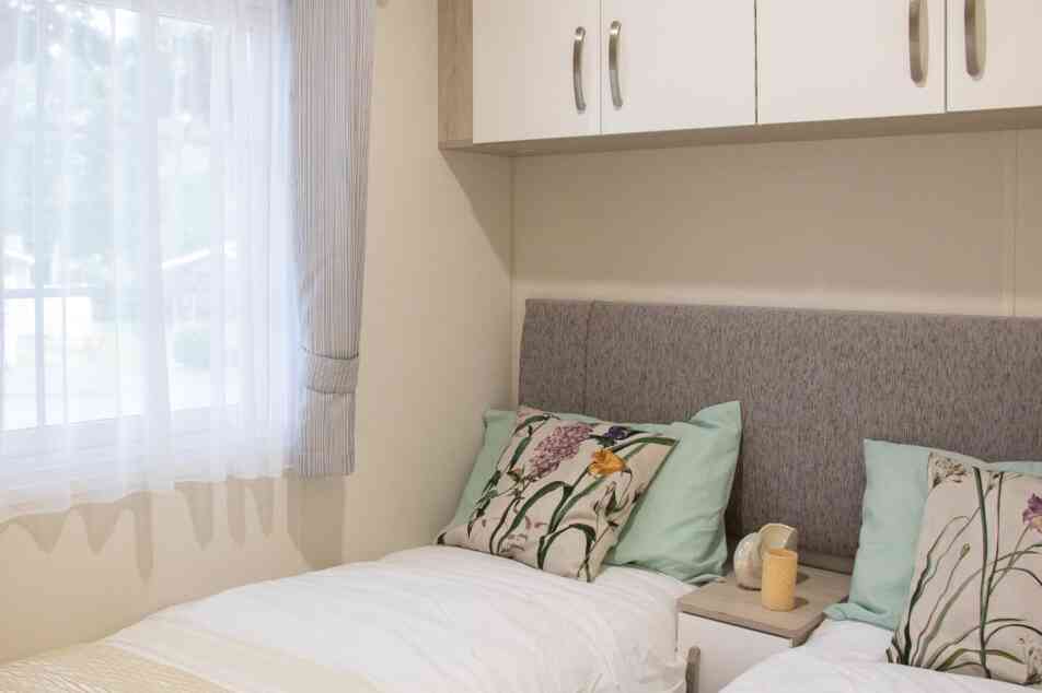 A cozy bedroom featuring two twin beds with light blue and floral-patterned pillows. There is a neutral-colored headboard and small bedside tables with decorative items. Natural light filters through a sheer window curtain.