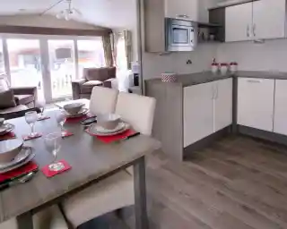 A modern kitchen and dining area featuring a wooden table set for six with plates, glasses, and napkins. The kitchen includes white cabinets, a microwave, and a countertop, with a living area visible in the background. Large windows allow natural light to fill the space.