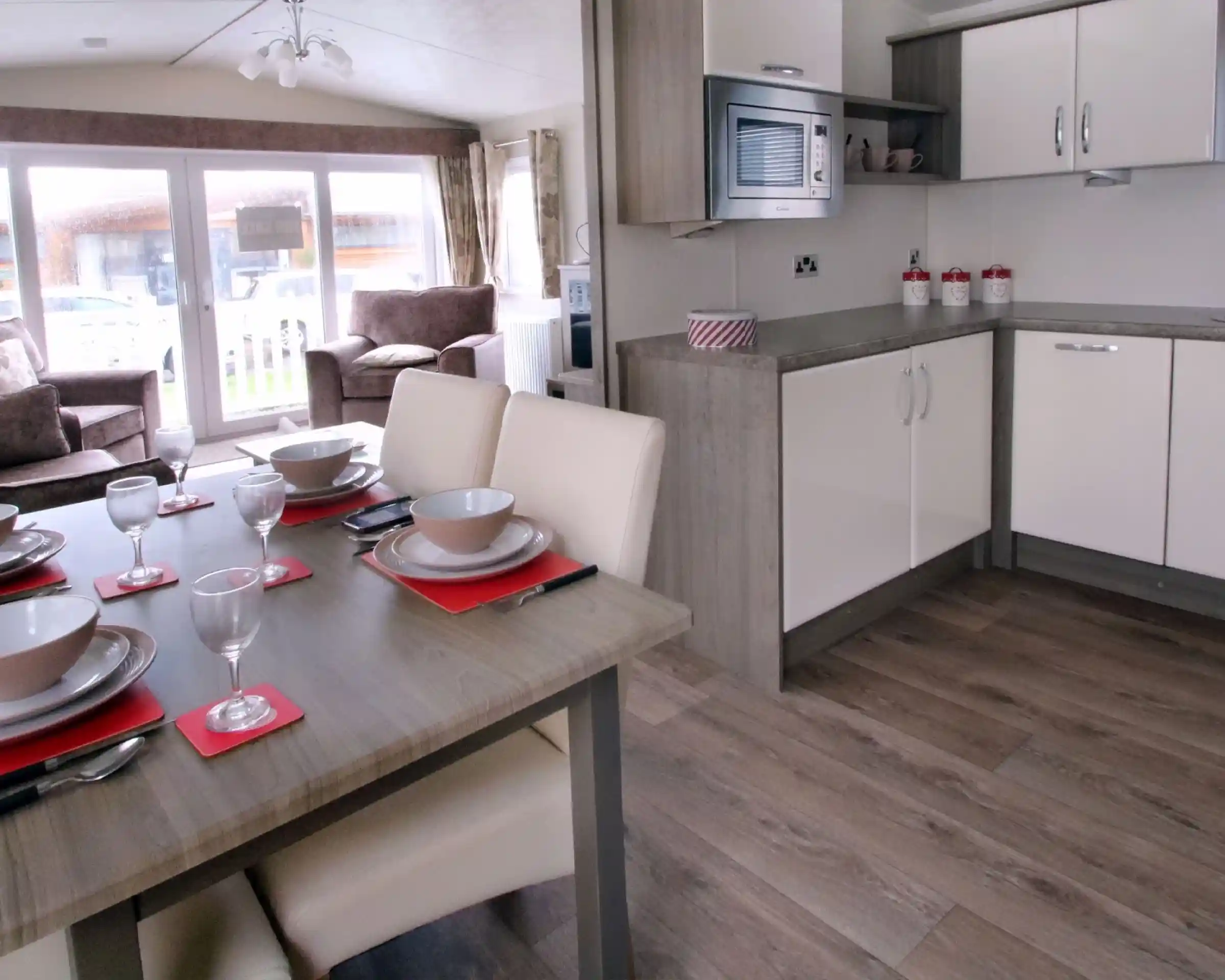 A modern kitchen and dining area featuring a wooden table set for six with plates, glasses, and napkins. The kitchen includes white cabinets, a microwave, and a countertop, with a living area visible in the background. Large windows allow natural light to fill the space.