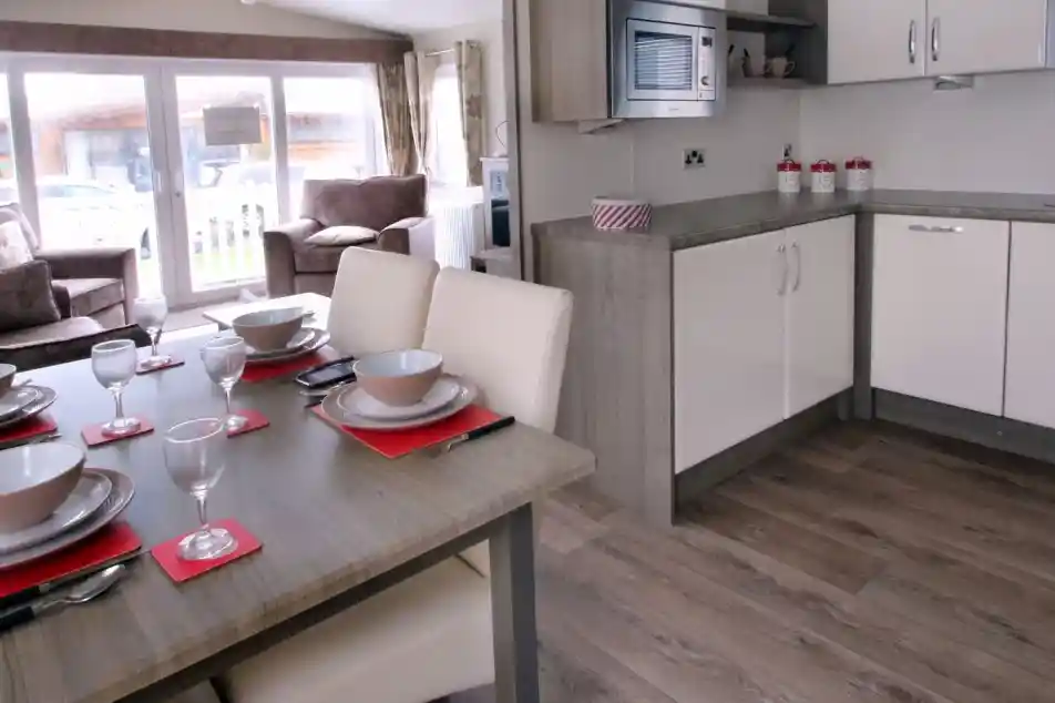 A modern kitchen and dining area featuring a wooden table set for six with plates, glasses, and napkins. The kitchen includes white cabinets, a microwave, and a countertop, with a living area visible in the background. Large windows allow natural light to fill the space.