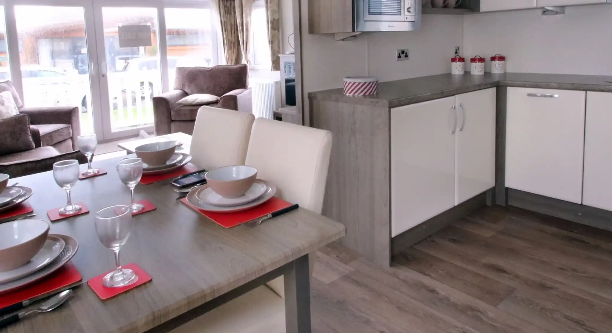 A modern kitchen and dining area featuring a wooden table set for six with plates, glasses, and napkins. The kitchen includes white cabinets, a microwave, and a countertop, with a living area visible in the background. Large windows allow natural light to fill the space.