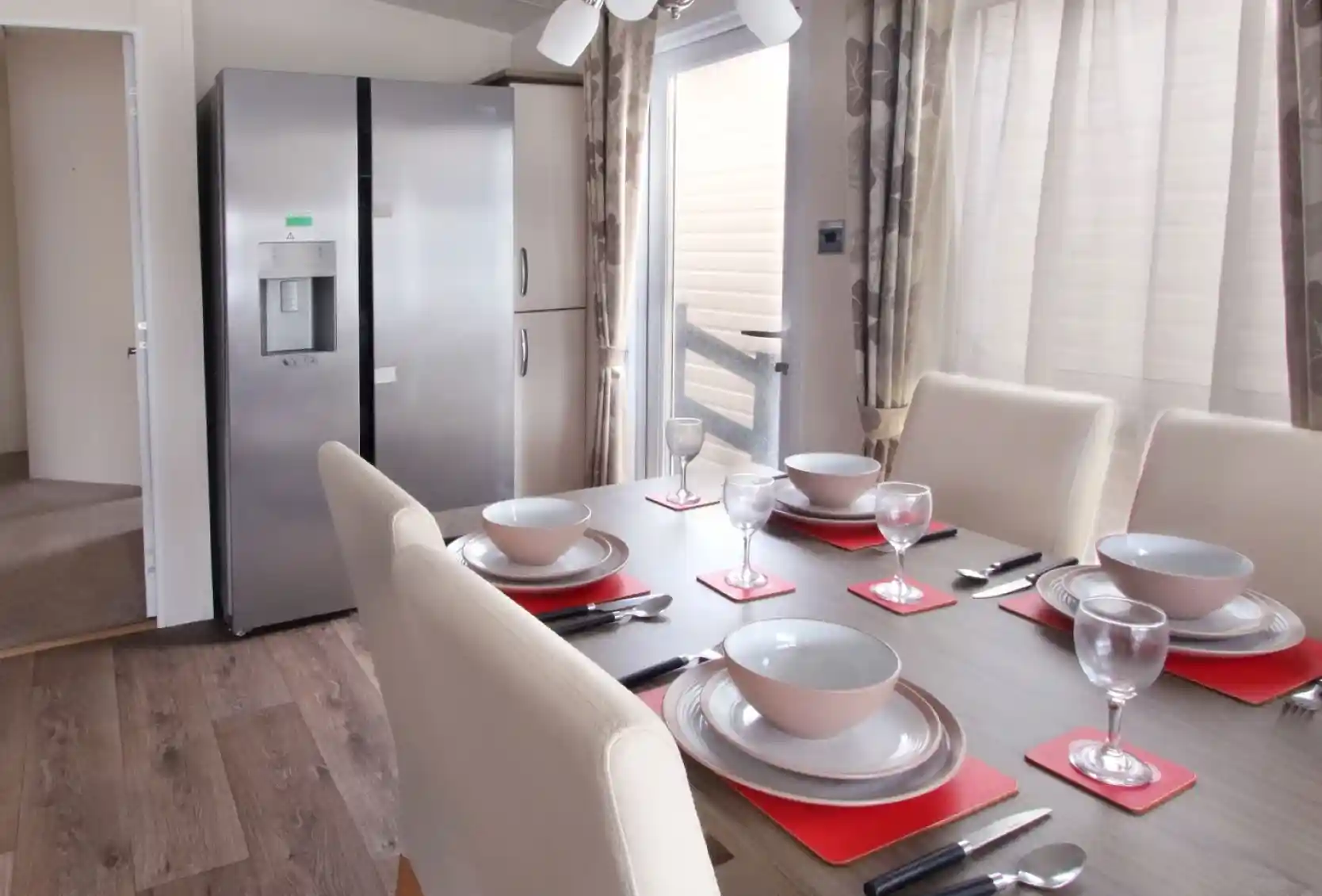 A modern dining area featuring a wooden table set with red placemats, bowls, and wine glasses. A large silver refrigerator stands nearby, with light streaming through a window adorned with sheer curtains.