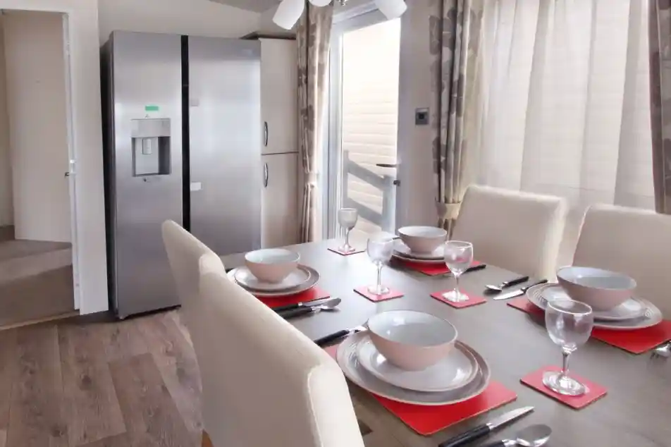 A modern dining area featuring a wooden table set with red placemats, bowls, and wine glasses. A large silver refrigerator stands nearby, with light streaming through a window adorned with sheer curtains.