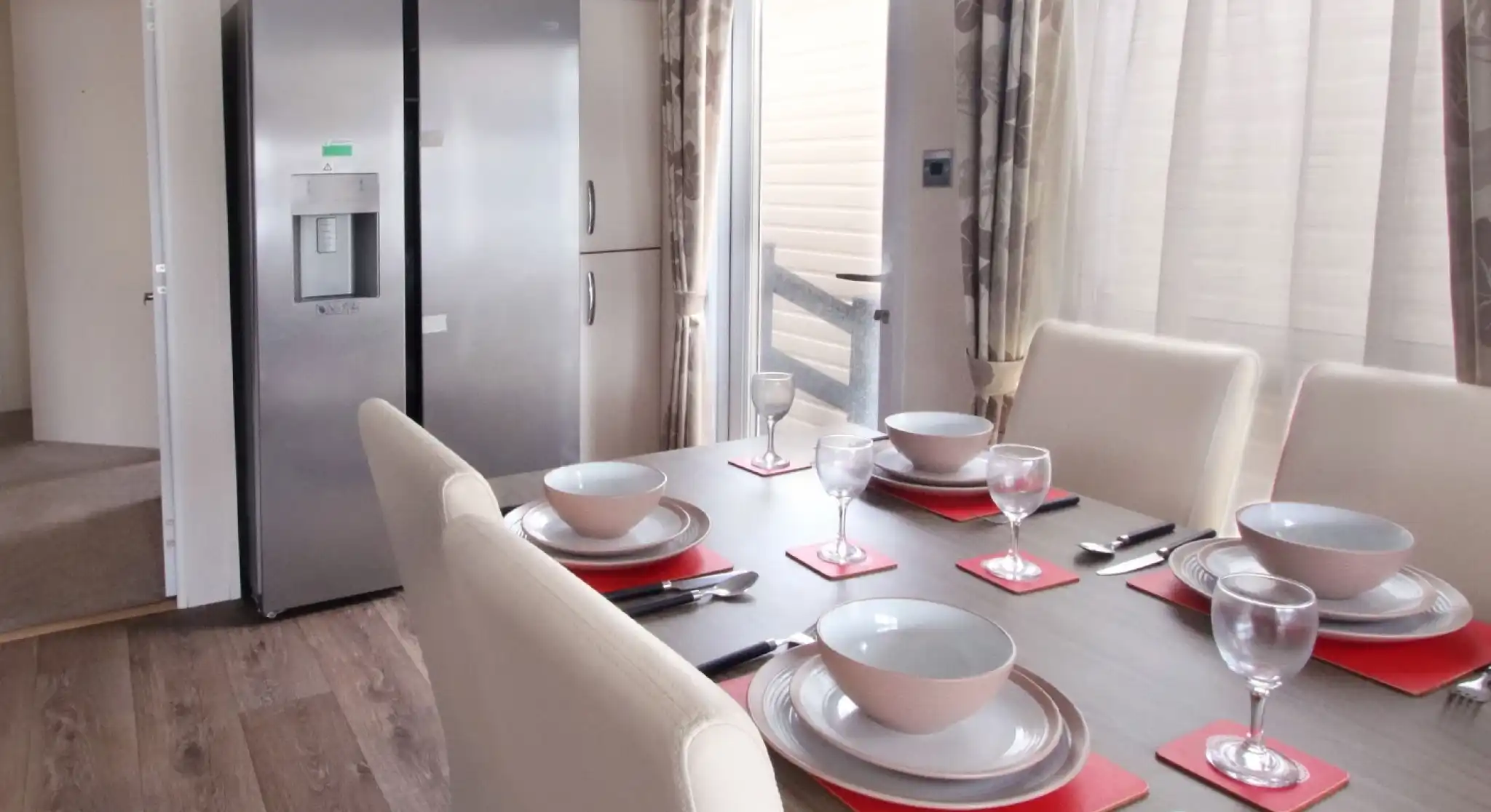 A modern dining area featuring a wooden table set with red placemats, bowls, and wine glasses. A large silver refrigerator stands nearby, with light streaming through a window adorned with sheer curtains.