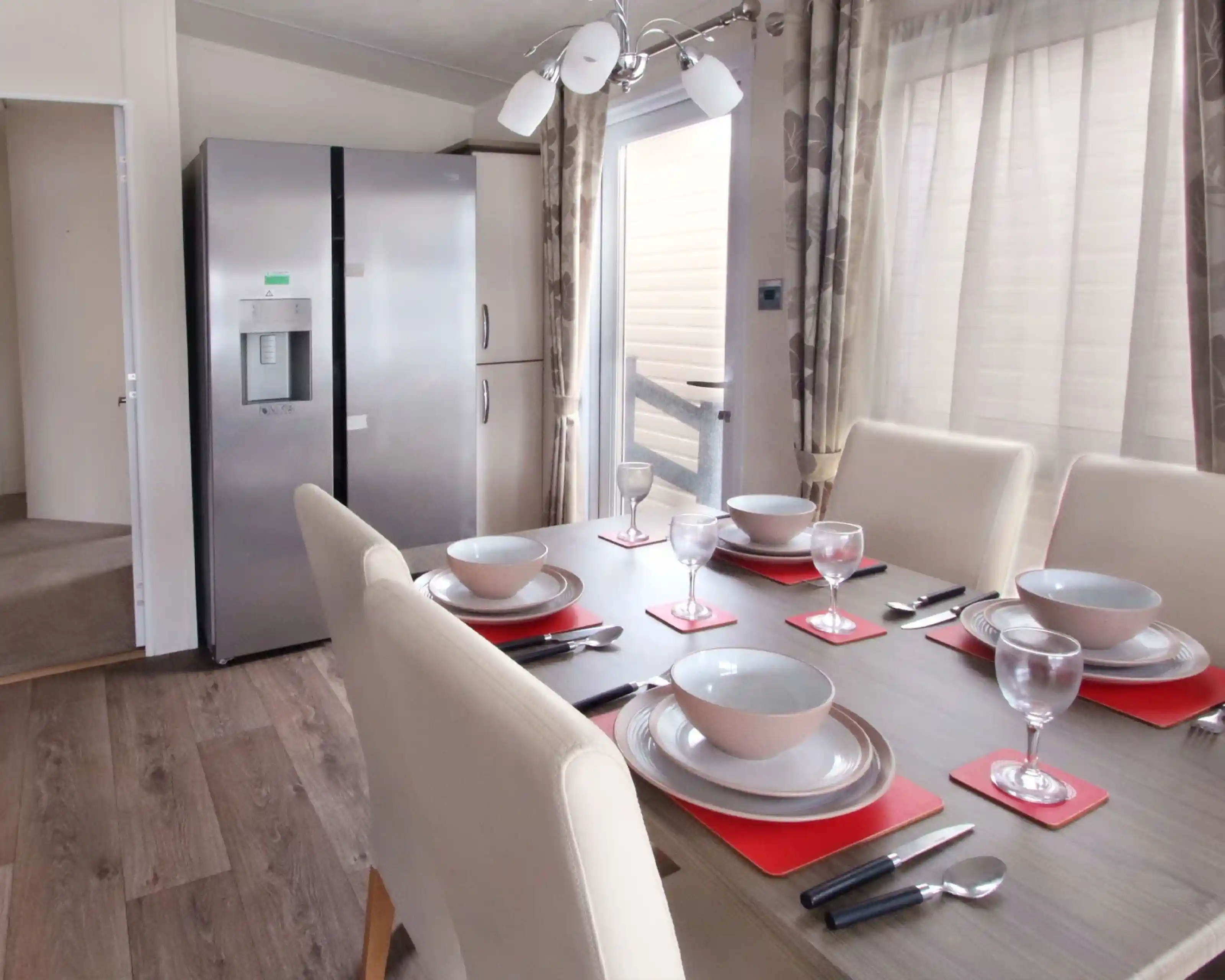 A modern dining area featuring a wooden table set with red placemats, bowls, and wine glasses. A large silver refrigerator stands nearby, with light streaming through a window adorned with sheer curtains.