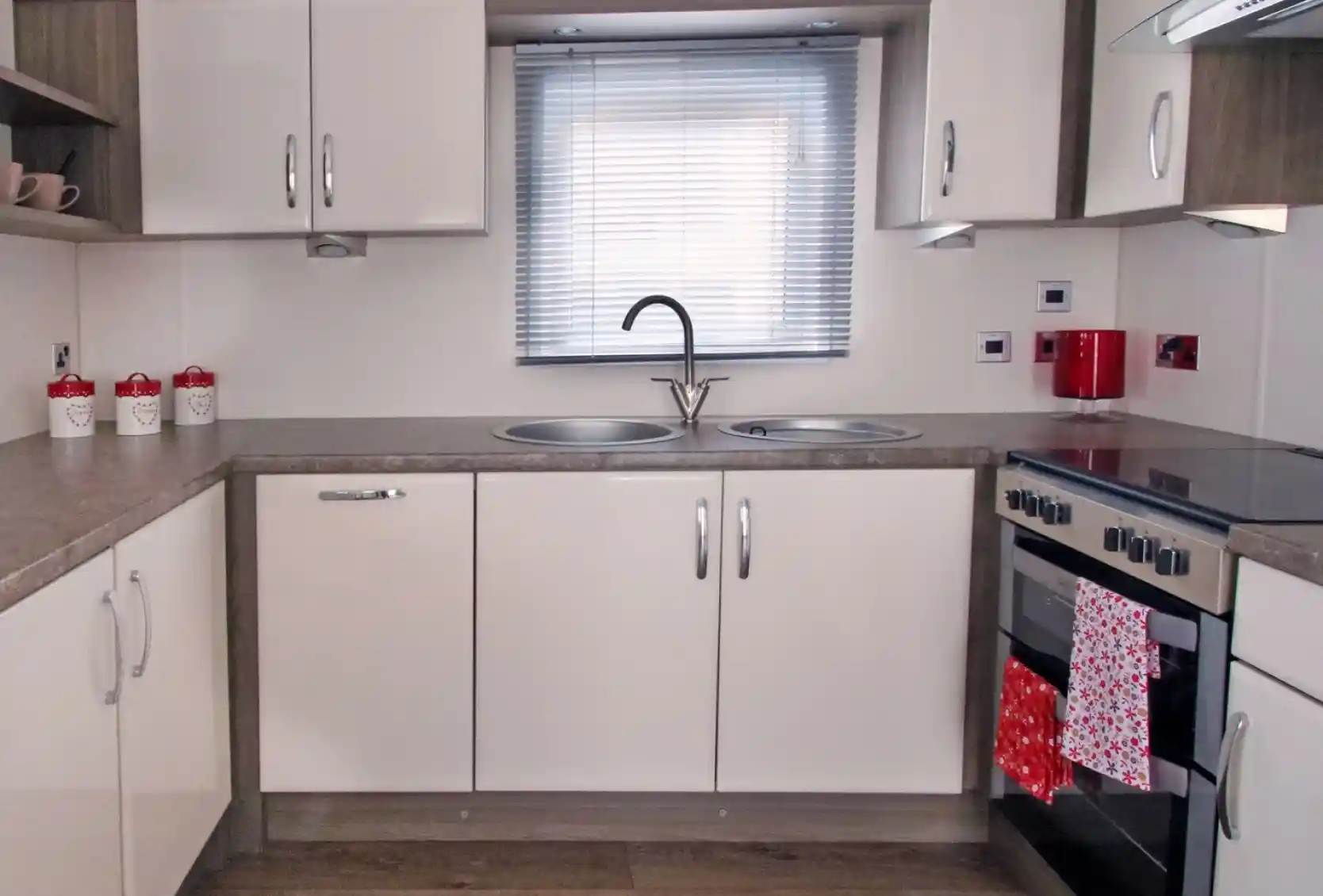 Modern kitchen with beige cabinets, gray countertops, a double sink under a window with blinds, and a stove with an oven. Red accents include a kettle and a towel hanging from the oven. Decorative jars are placed on the shelf.