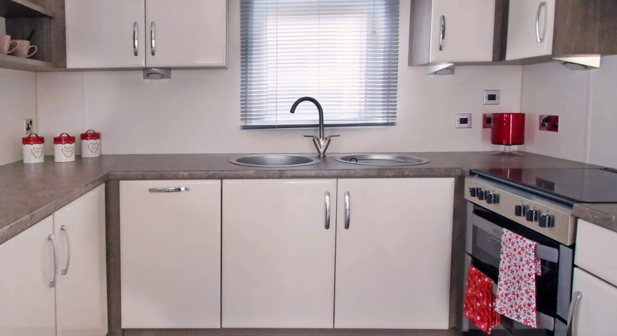 Modern kitchen with beige cabinets, gray countertops, a double sink under a window with blinds, and a stove with an oven. Red accents include a kettle and a towel hanging from the oven. Decorative jars are placed on the shelf.