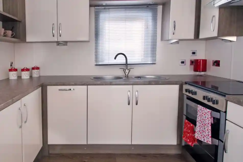 Modern kitchen with beige cabinets, gray countertops, a double sink under a window with blinds, and a stove with an oven. Red accents include a kettle and a towel hanging from the oven. Decorative jars are placed on the shelf.