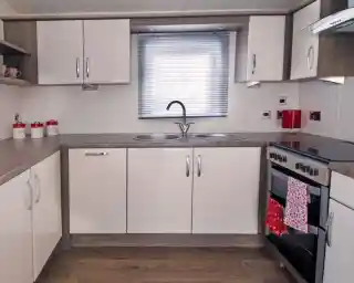 Modern kitchen with beige cabinets, gray countertops, a double sink under a window with blinds, and a stove with an oven. Red accents include a kettle and a towel hanging from the oven. Decorative jars are placed on the shelf.