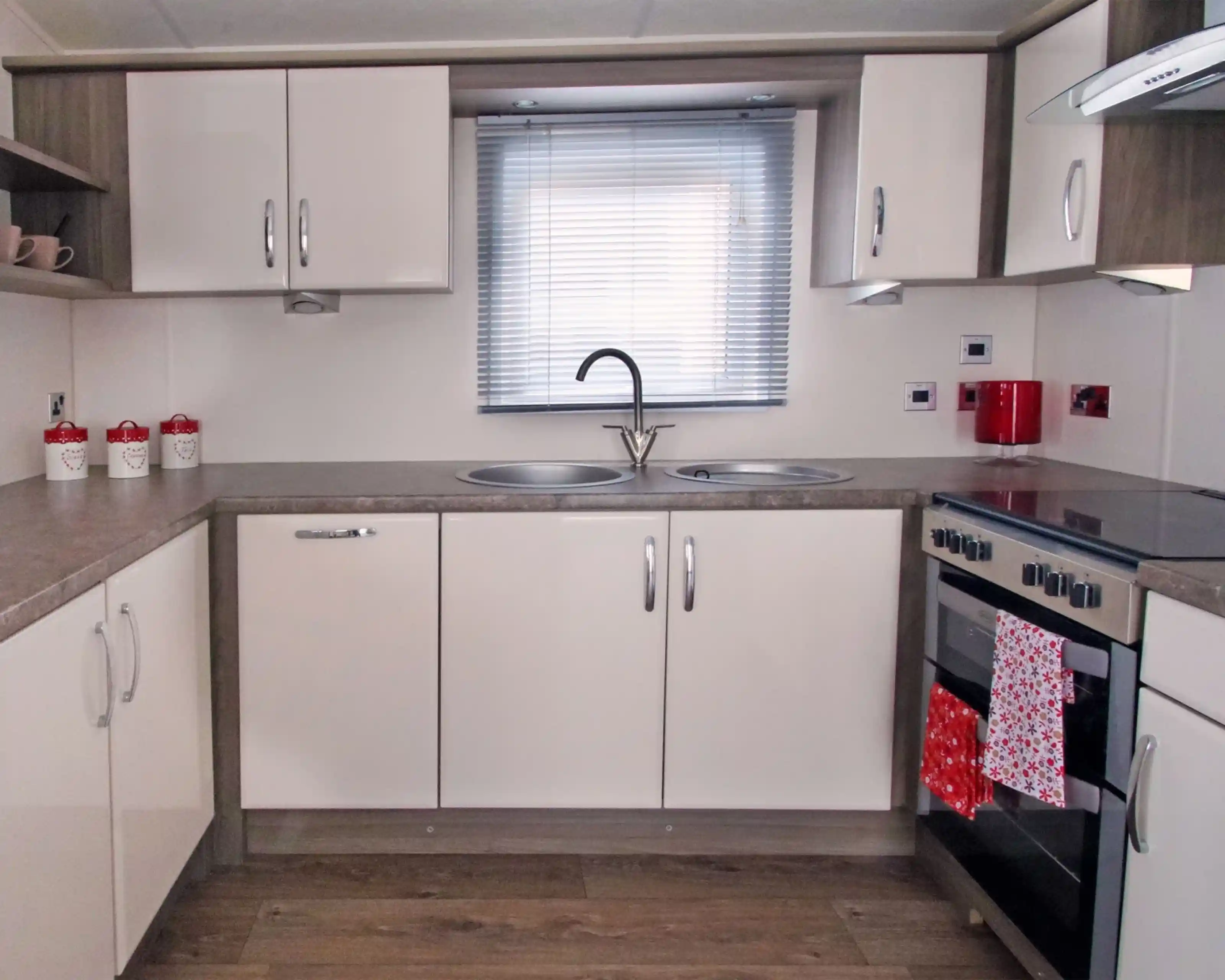 Modern kitchen with beige cabinets, gray countertops, a double sink under a window with blinds, and a stove with an oven. Red accents include a kettle and a towel hanging from the oven. Decorative jars are placed on the shelf.