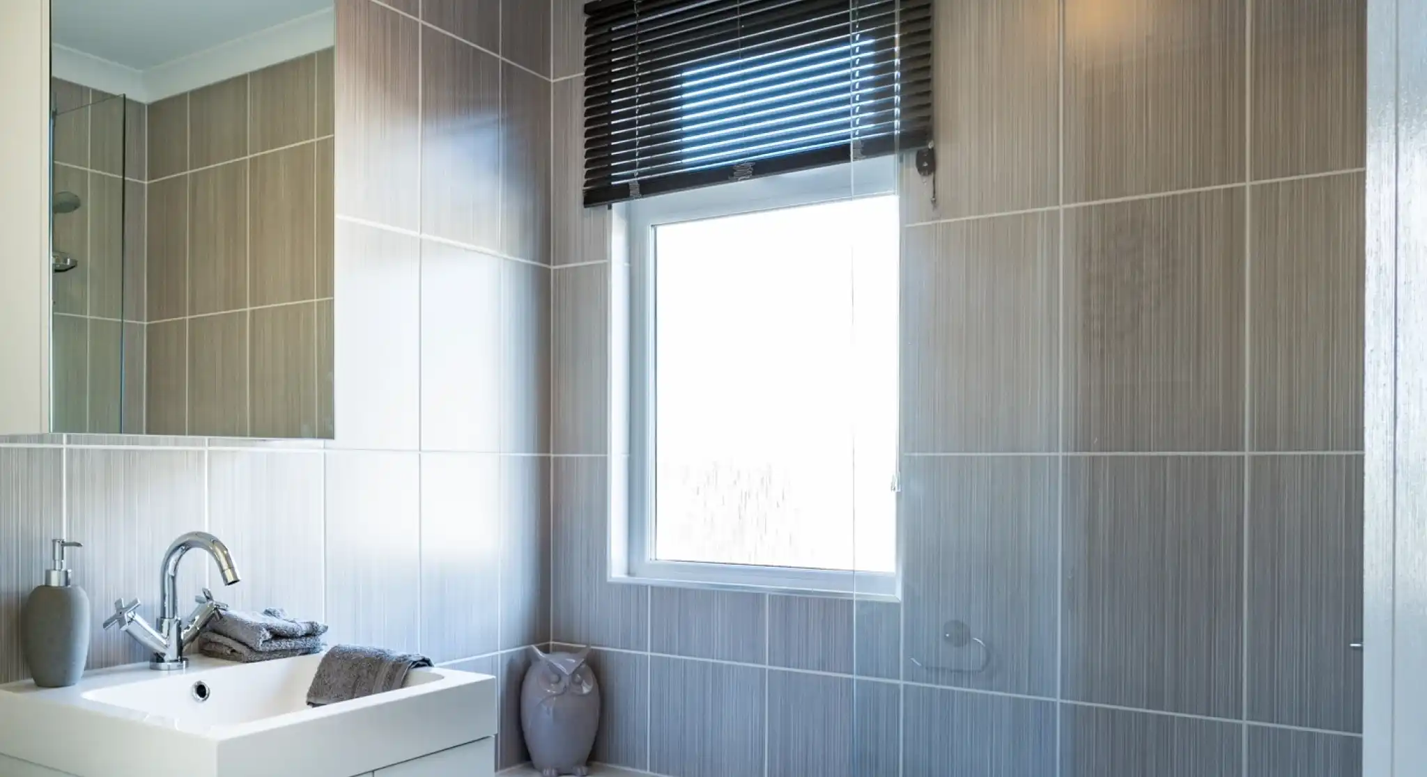 A bathroom with white gloss units including a vanity unit with inset wash basin, a bath with shower screen and shower over and a mirrored cabinet. A window with blind allows in natural light.