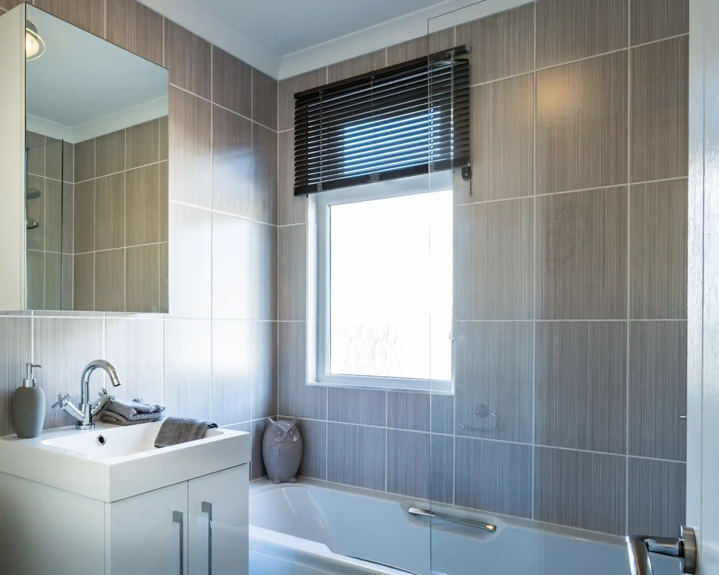 A bathroom with white gloss units including a vanity unit with inset wash basin, a bath with shower screen and shower over and a mirrored cabinet. A window with blind allows in natural light.