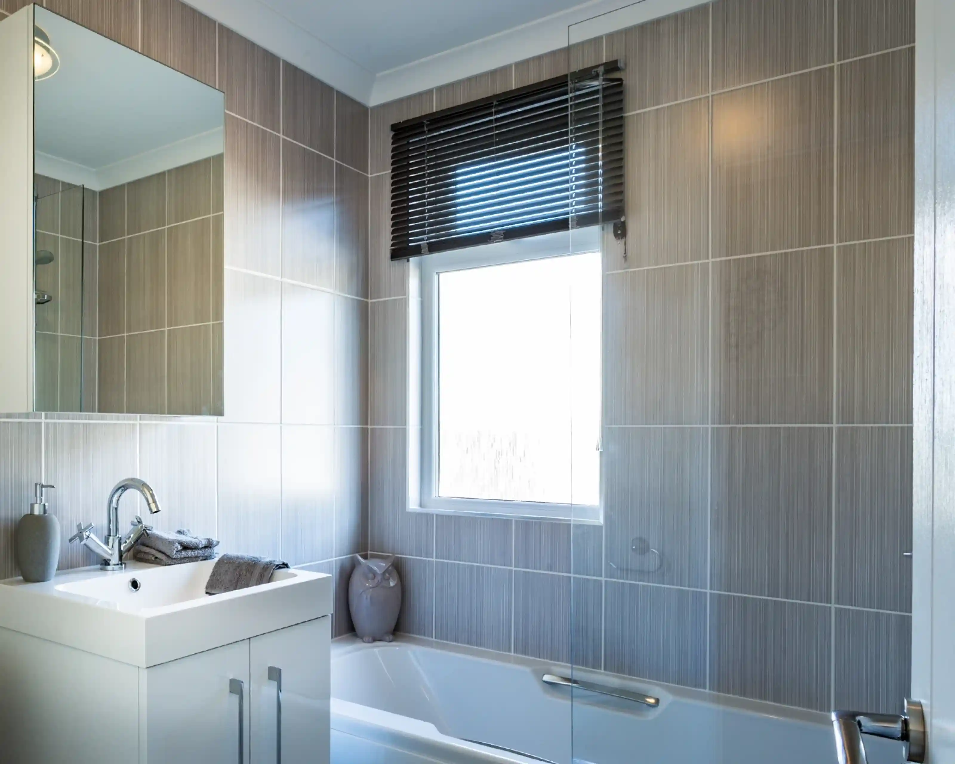 A bathroom with white gloss units including a vanity unit with inset wash basin, a bath with shower screen and shower over and a mirrored cabinet. A window with blind allows in natural light.