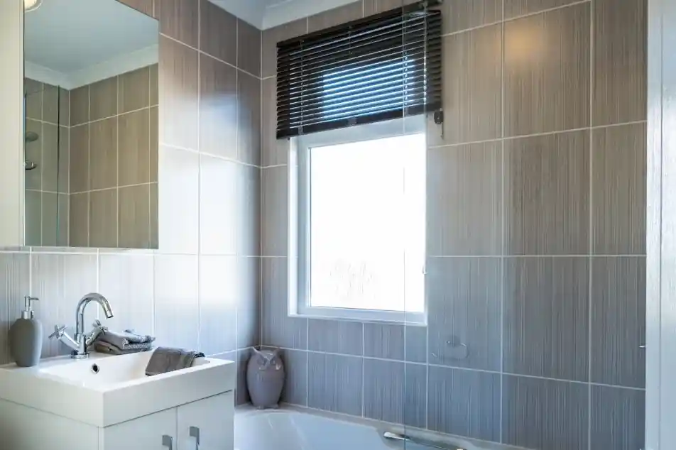 A bathroom with white gloss units including a vanity unit with inset wash basin, a bath with shower screen and shower over and a mirrored cabinet. A window with blind allows in natural light.