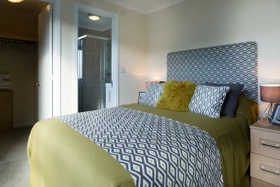 A master bedroom featuring a double bed with patterned yellow and grey headboard, cushions and bedspread. There is an en suite shower room and a walk-in wardrobe together with bedside tables and lamps.