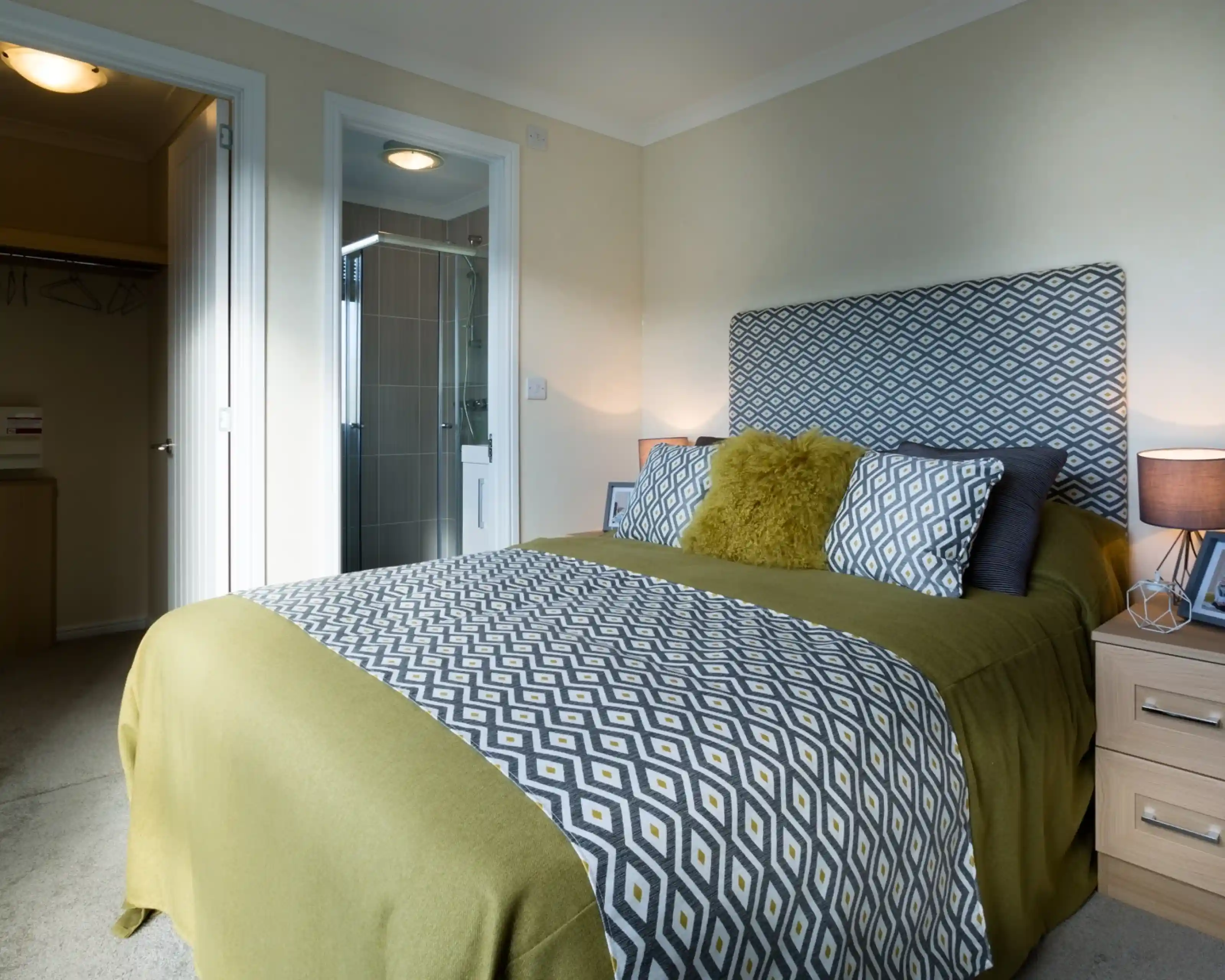 A master bedroom featuring a double bed with patterned yellow and grey headboard, cushions and bedspread. There is an en suite shower room and a walk-in wardrobe together with bedside tables and lamps.