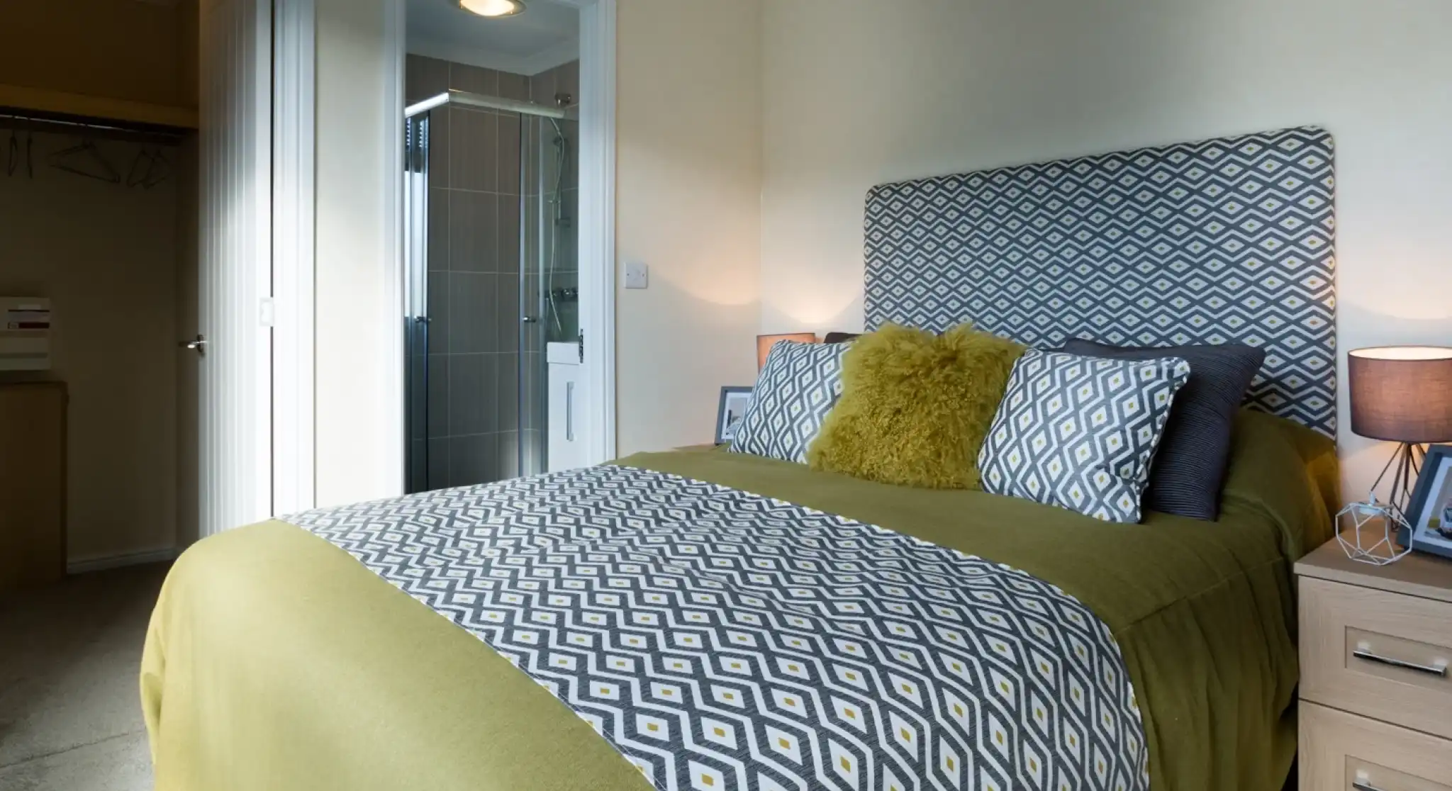 A master bedroom featuring a double bed with patterned yellow and grey headboard, cushions and bedspread. There is an en suite shower room and a walk-in wardrobe together with bedside tables and lamps.